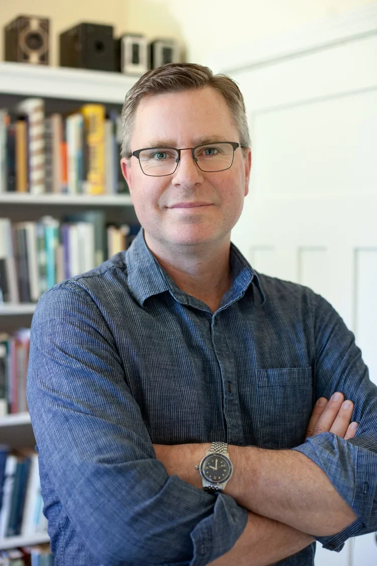 Dominic Smith, a man with glasses and a blue shirt crossing his arms in front of a bookshelf filled with books, in a well-lit room.
