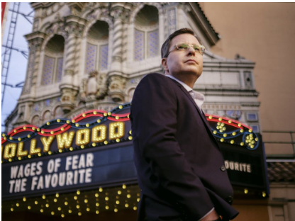 A man wearing glasses and a dark suit stands outside a historic theater with a marquee displaying 'Wages of Fear, The Favourite,' in front of the Hollywood Theatre in Los Angeles.