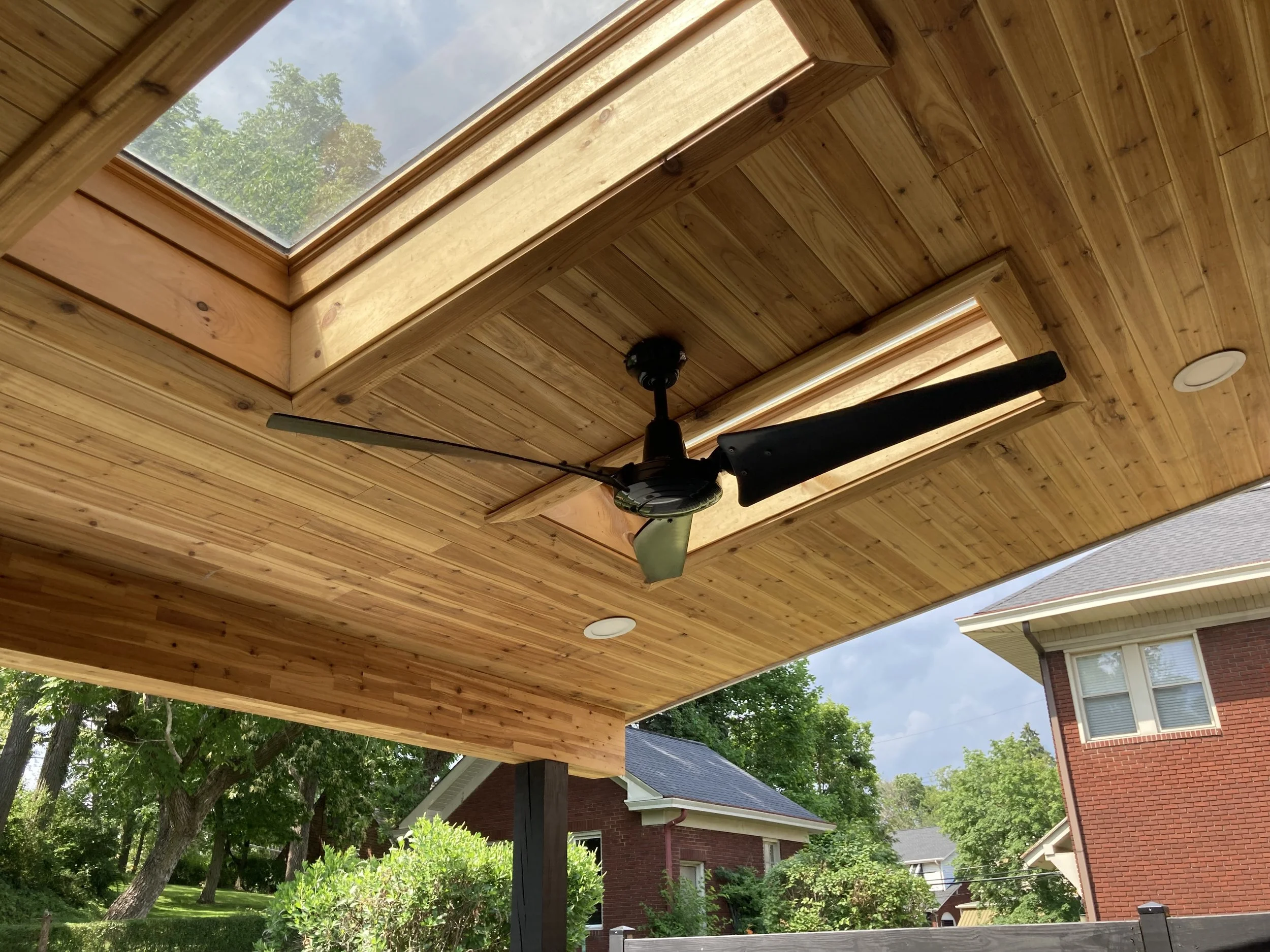 Under a wooden porch ceiling, a black ceiling fan with three blades is installed. The ceiling has two skylights, and outside there are trees and houses with brick exteriors.
