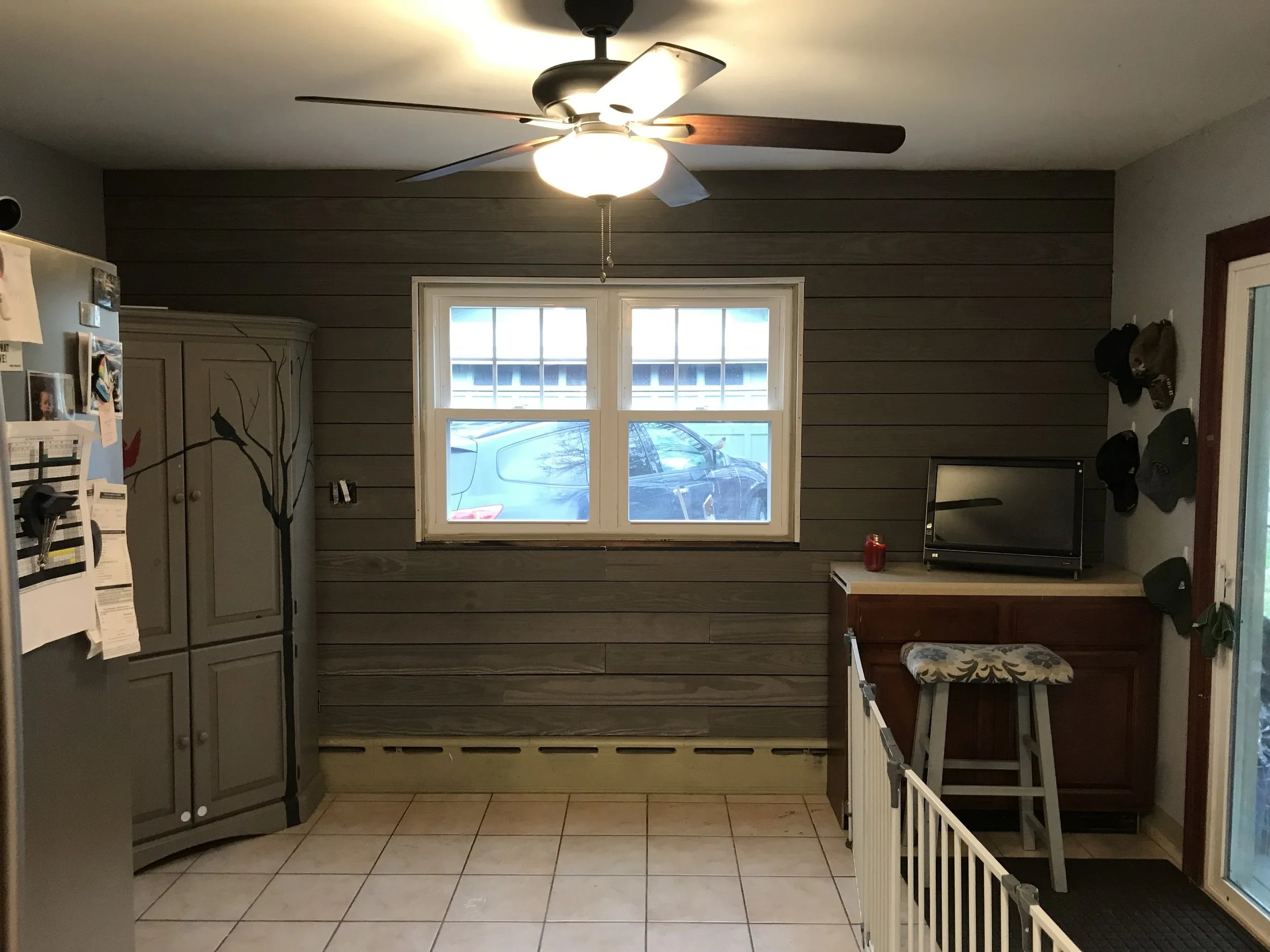 Interior of a kitchen with a gray fridge covered with papers on the left, a decorative gray cabinet with a branch motif, a window showing a parked car outside, a ceiling fan with lights, and a small TV on a wooden counter with hats on the wall nearby.
