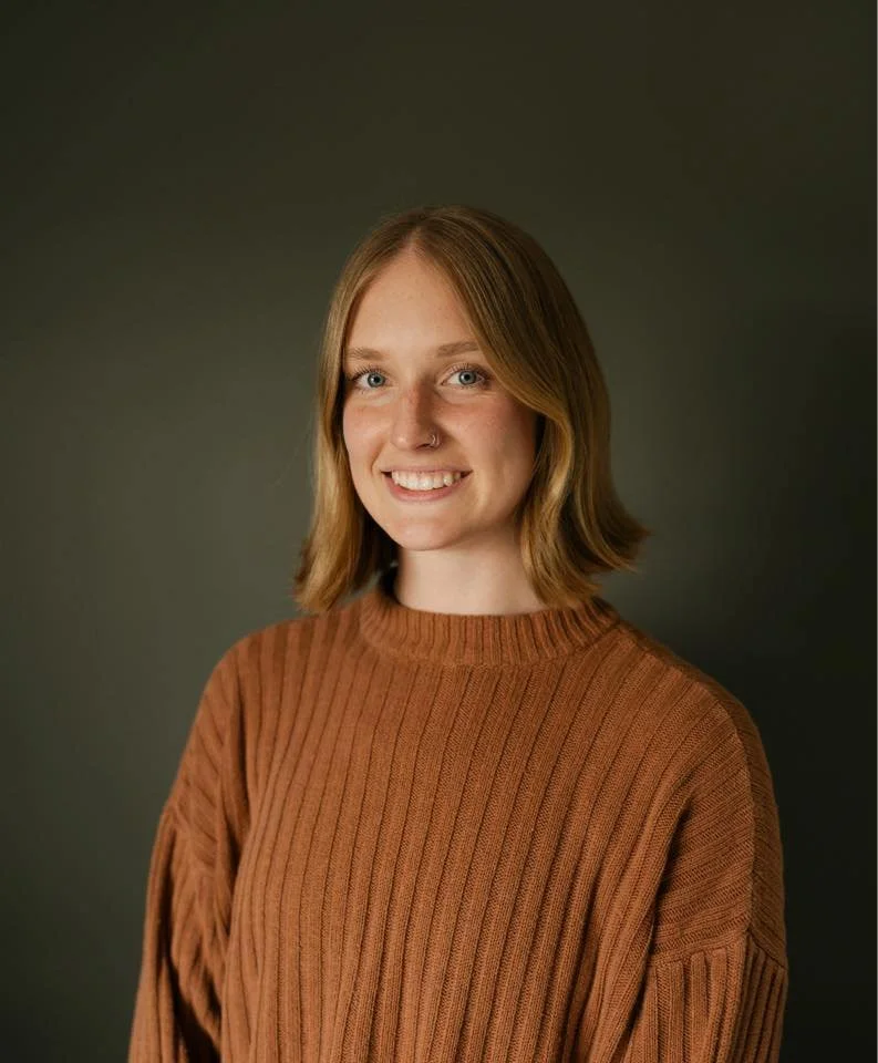 Portrait of a young woman with shoulder-length light brown hair, wearing a brown ribbed sweater, smiling against a dark background.