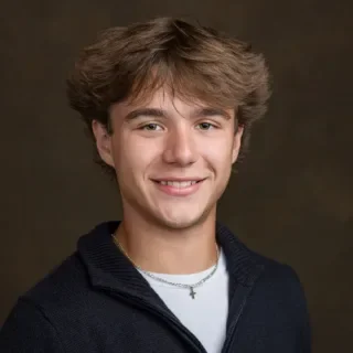 A young man with brown hair and a smile, wearing a dark sweater and a necklace, against a plain background.