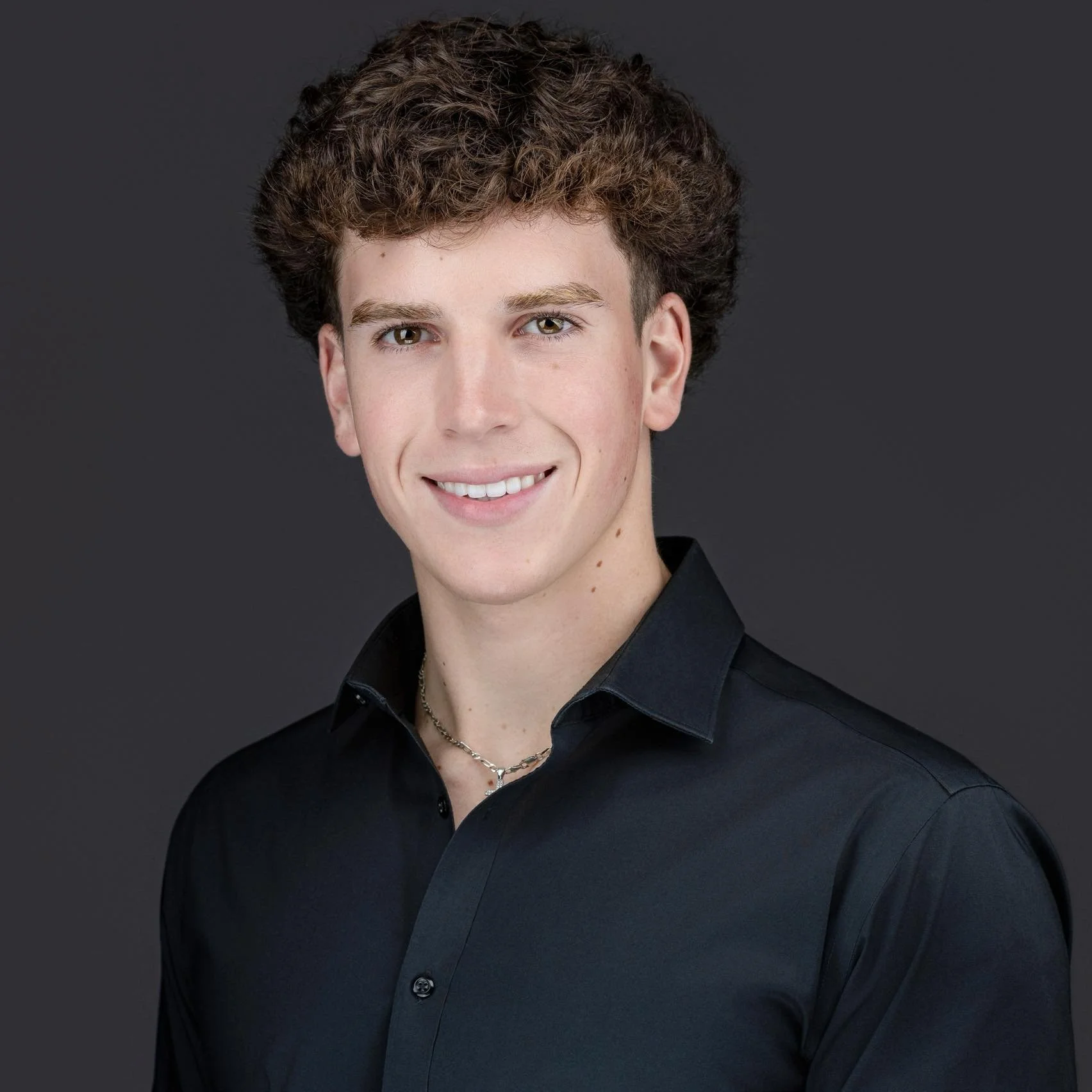 A young man with curly brown hair and light skin, smiling while wearing a black collared shirt and a silver necklace, against a dark gray background.