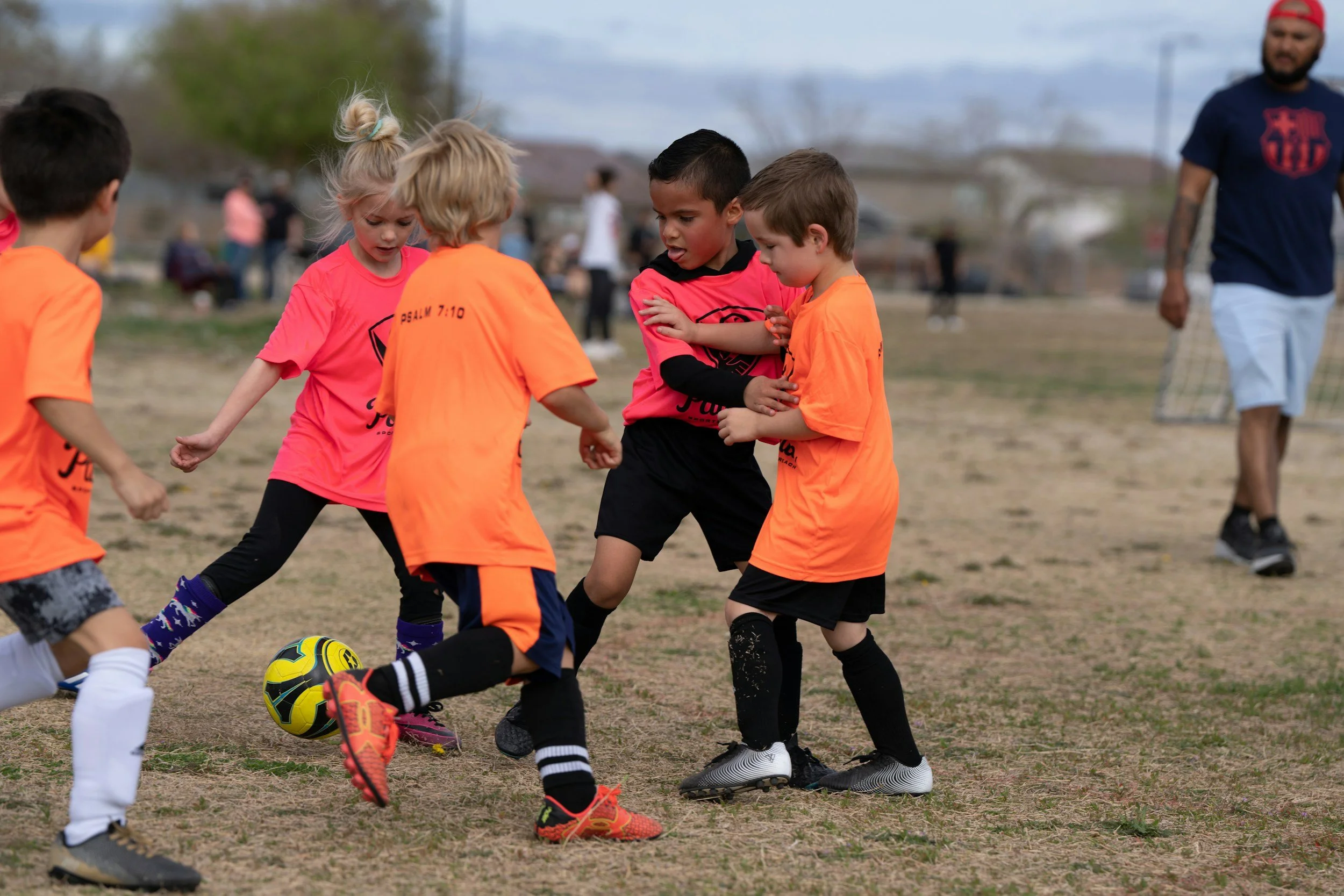 Children in playing soccer on a field, with a ball near their feet, and a coach walking in the background.