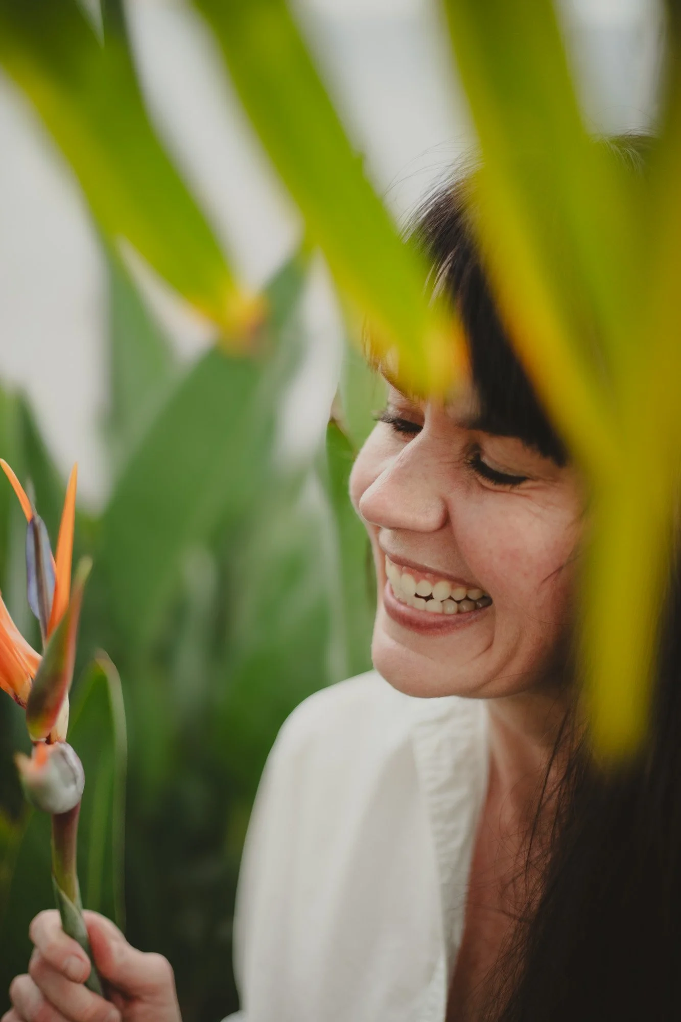 A woman smiling and holding a flower, surrounded by green leaves.