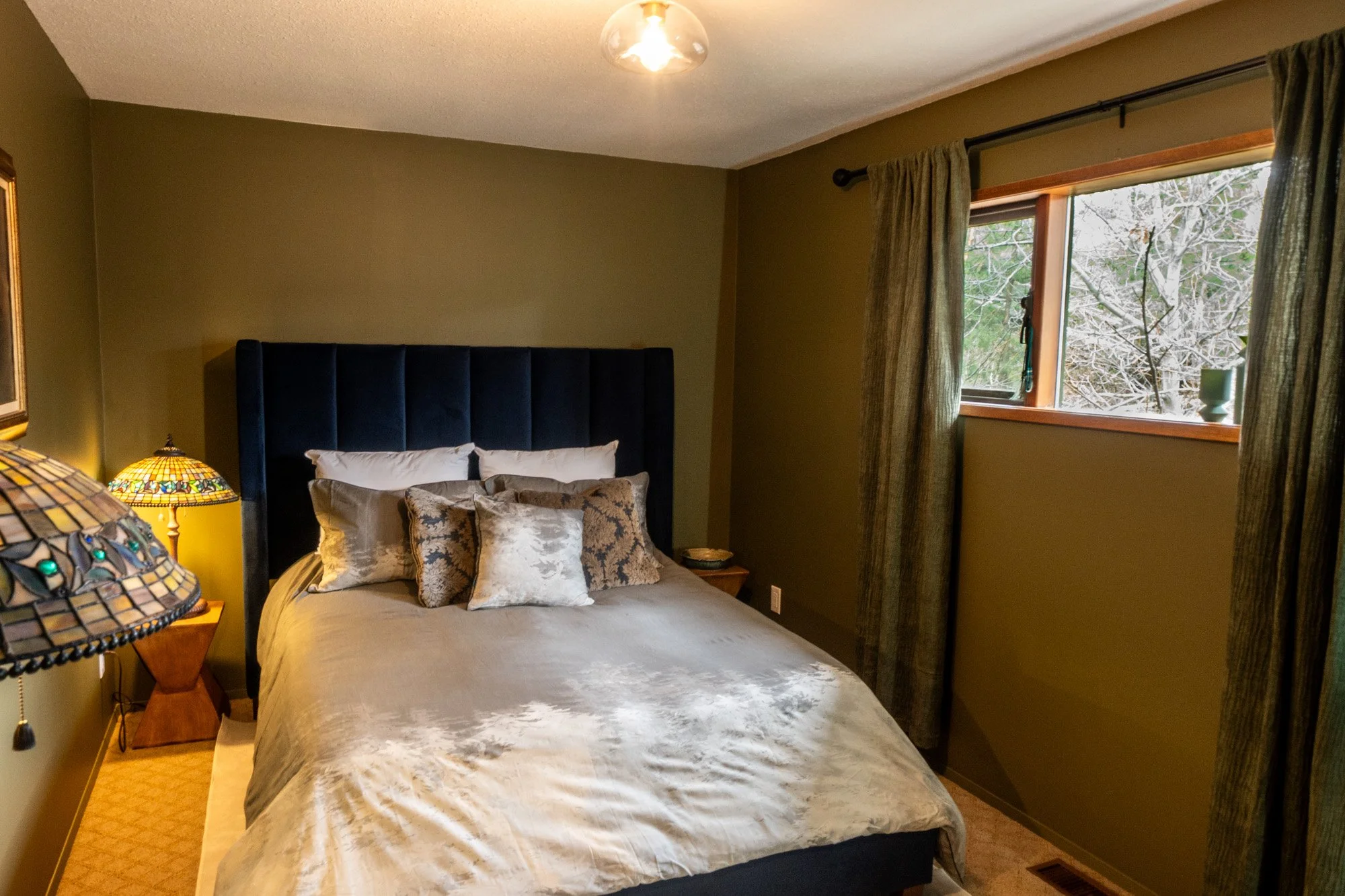 A bedroom with a bed featuring a large blue headboard, multiple pillows, and a beige bedspread. There is a window with green curtains on the right side showing a view of snow-covered trees. A Tiffany-style table lamp is on a wooden nightstand on the left side of the bed.