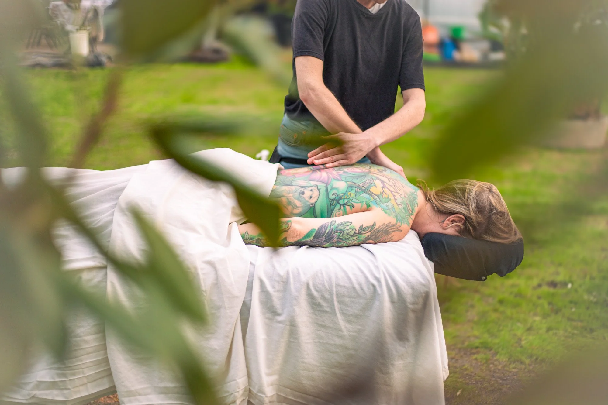 A person receiving an outdoor massage while lying face down on a massage table with a white sheet, amidst greenery and blurred leaves in the foreground.