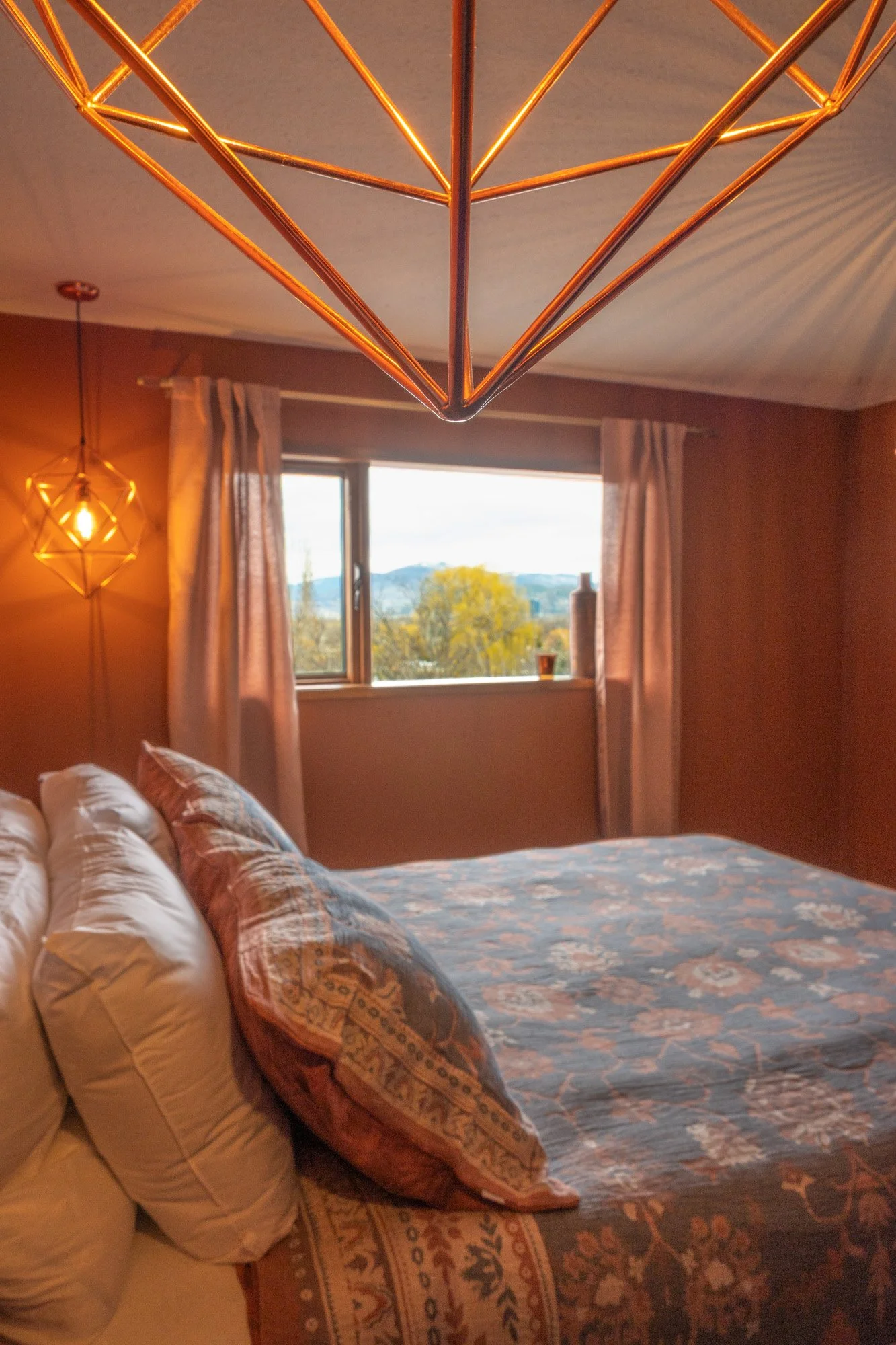 A bedroom with a bed made up with patterned bedding and pillows, a window with a view of trees and hills, two geometric pendant lights hanging from the ceiling, and curtains on either side of the window.