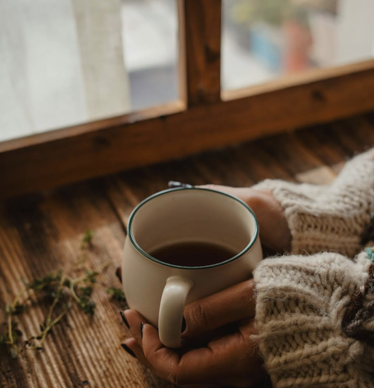 Person holding a white mug with a green rim, filled with a dark beverage, near a window with wooden framing, on a wooden table, wearing a chunky knit sweater.