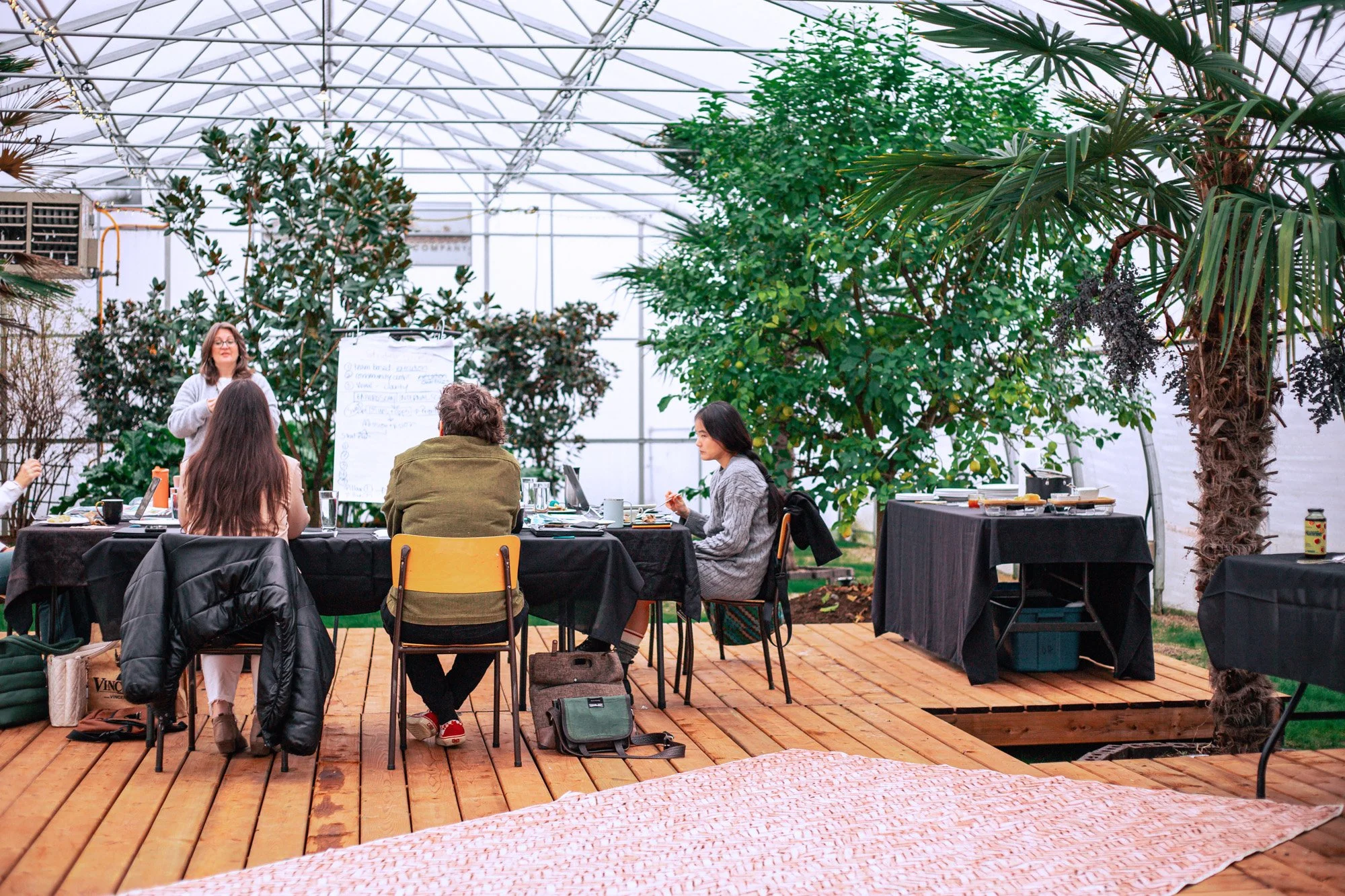 A group of people attending a presentation or meeting inside a greenhouse, surrounded by lush green plants and trees, with a woman standing near a whiteboard.