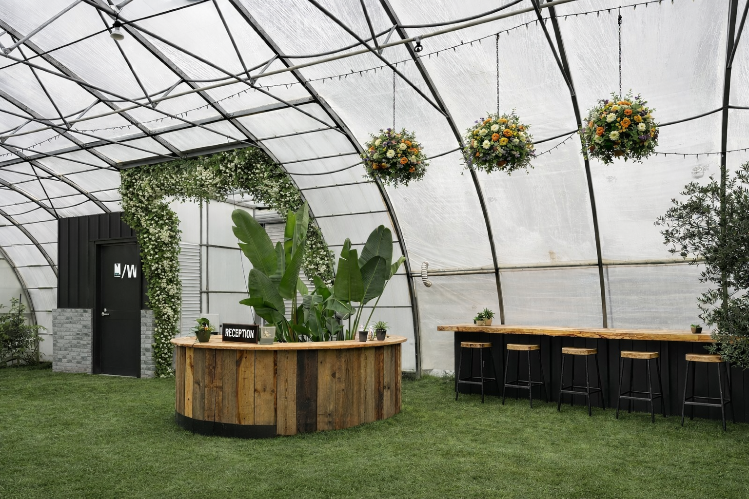 Indoor event space with floral decorations, reception desk, bar-height seating area, plants, and a curved ceiling structure.
