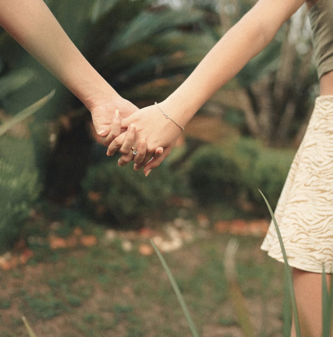 Two people holding hands outdoors, with a blurred green leafy background.