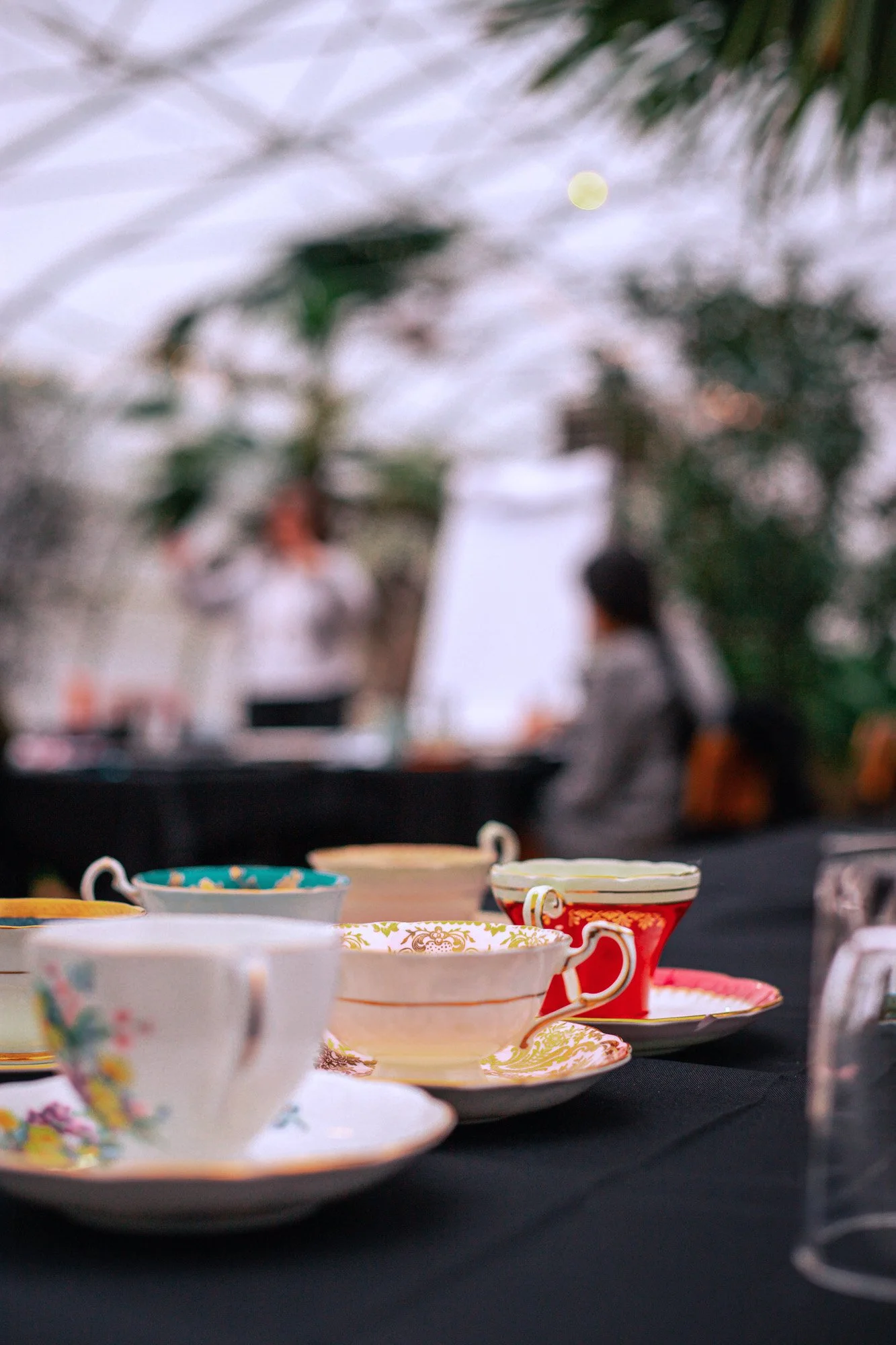 A table with colorful, patterned teacups and saucers on a dark tablecloth, with people and plants blurred in the background.