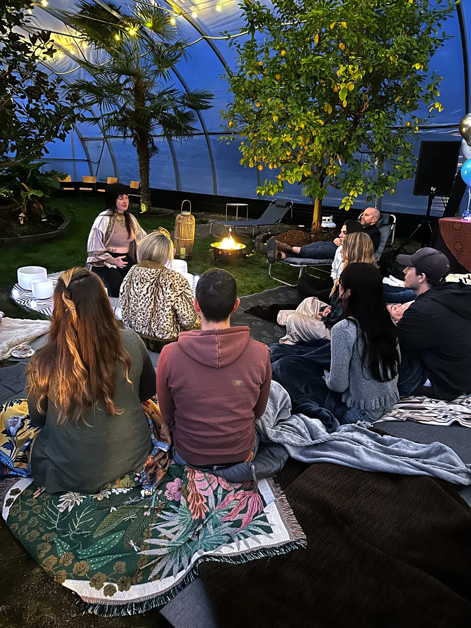 A group of people sitting on blankets and pillows in a semi-circle inside a glasshouse with lush green plants, trees, and string lights, attending a live outdoor performance near a fire pit.
