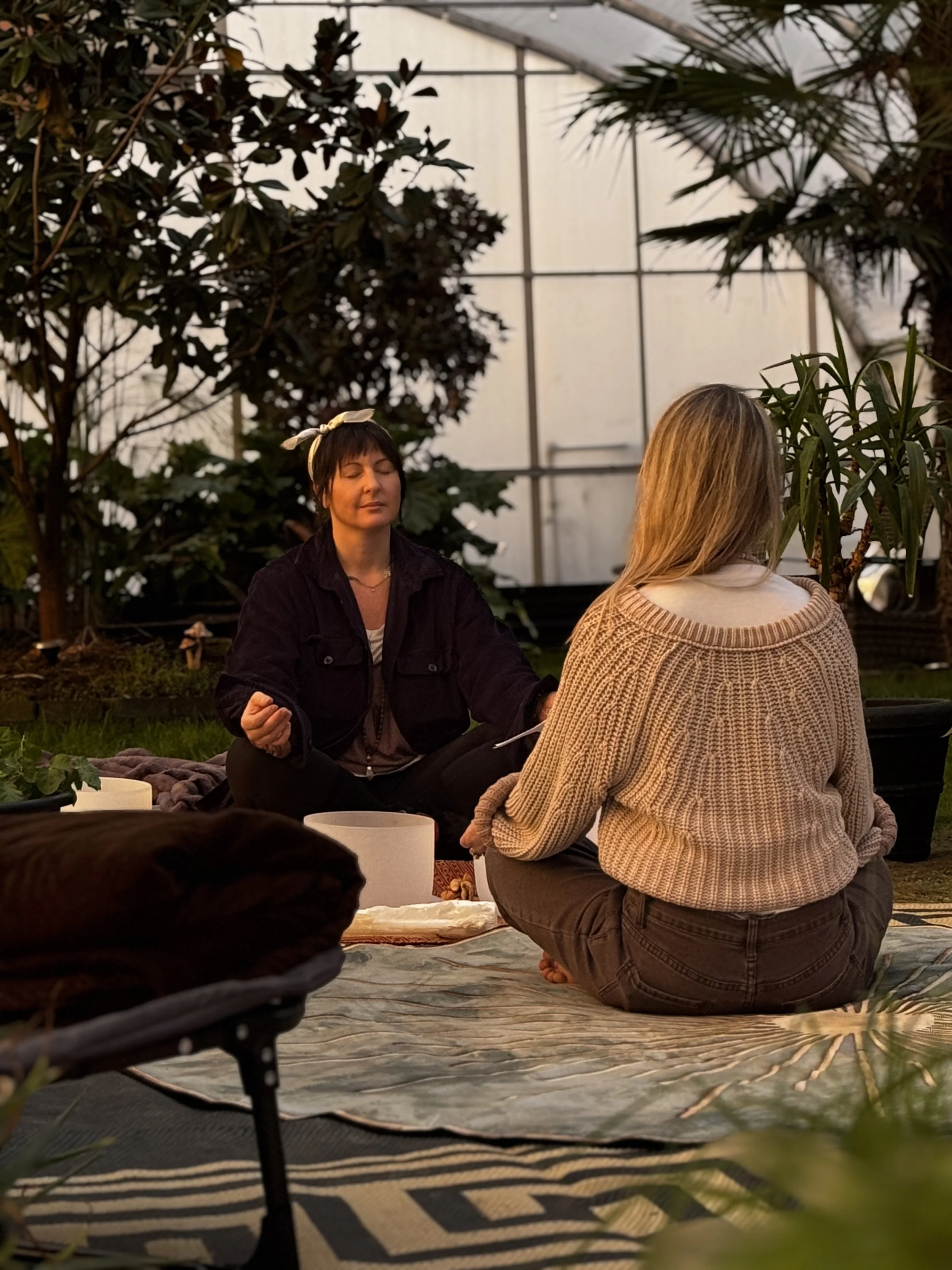 Two women sitting cross-legged on a blanket on the ground in a greenhouse, engaging in meditation or a spiritual practice, with plants and trees surrounding them.