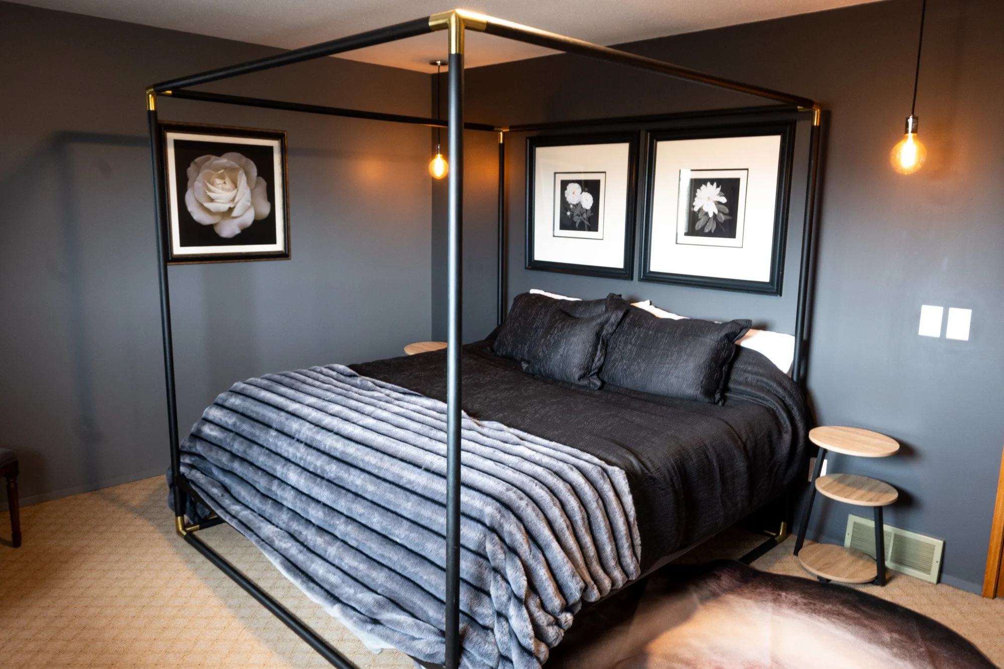 Modern bedroom featuring a black four-poster bed with black bedding and pillows, dark gray walls, three framed floral pictures above the bed, hanging Edison bulbs, and a small three-tier wood side table.