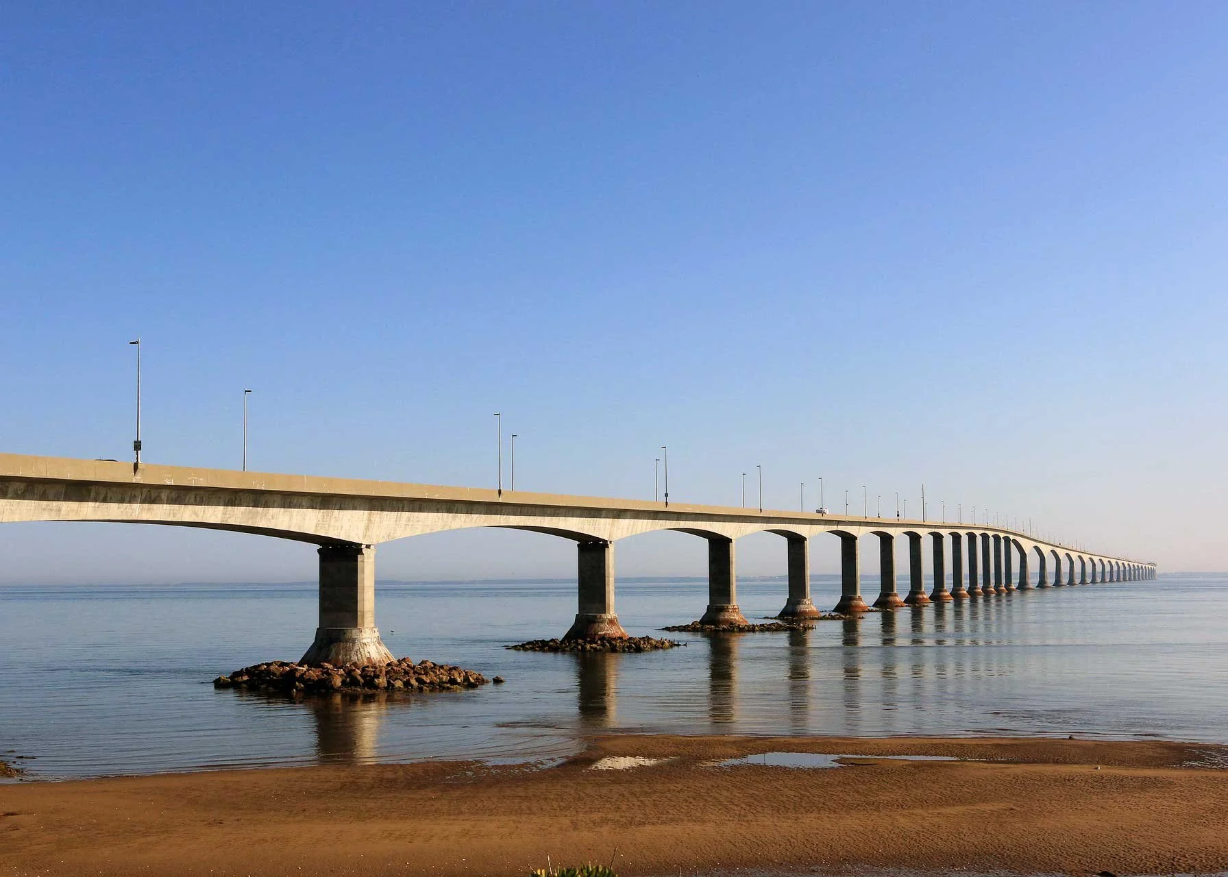 A long concrete bridge extending over calm water with a sandy shore in the foreground and a clear blue sky above.