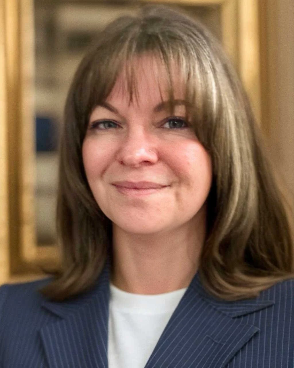 Close-up portrait of a woman with shoulder-length brown hair and light skin, wearing a navy pinstripe blazer and a white shirt.
