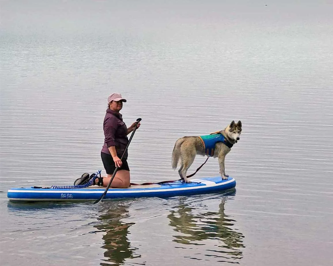 A woman kneeling on a paddleboard with a dog standing next to her on the same paddleboard, both on a calm body of water.