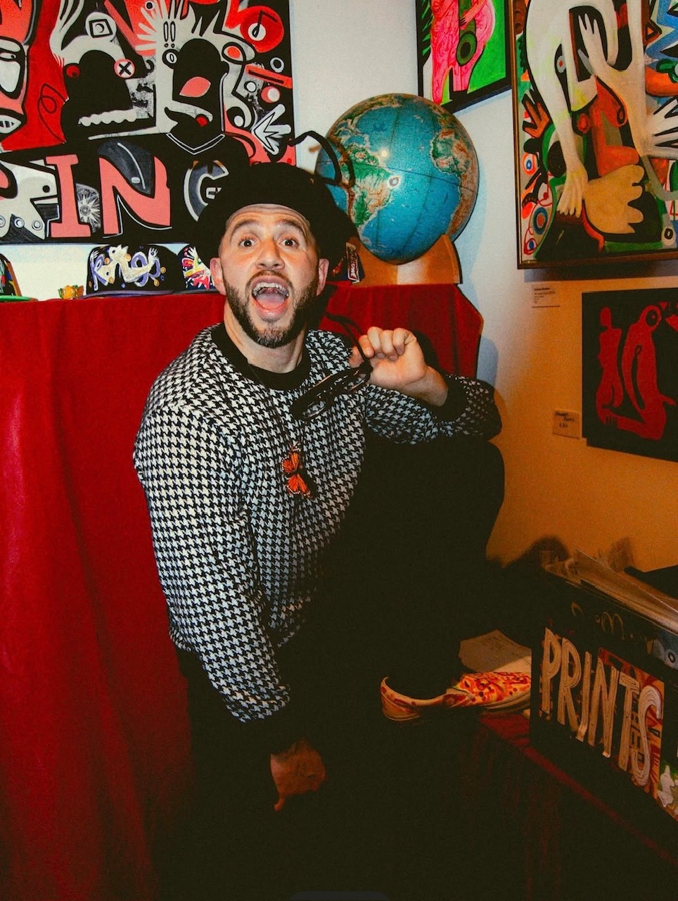 Man with a beard, wearing a black and white houndstooth sweater, holding a pair of glasses, in an art gallery with colorful abstract paintings and a globe background.