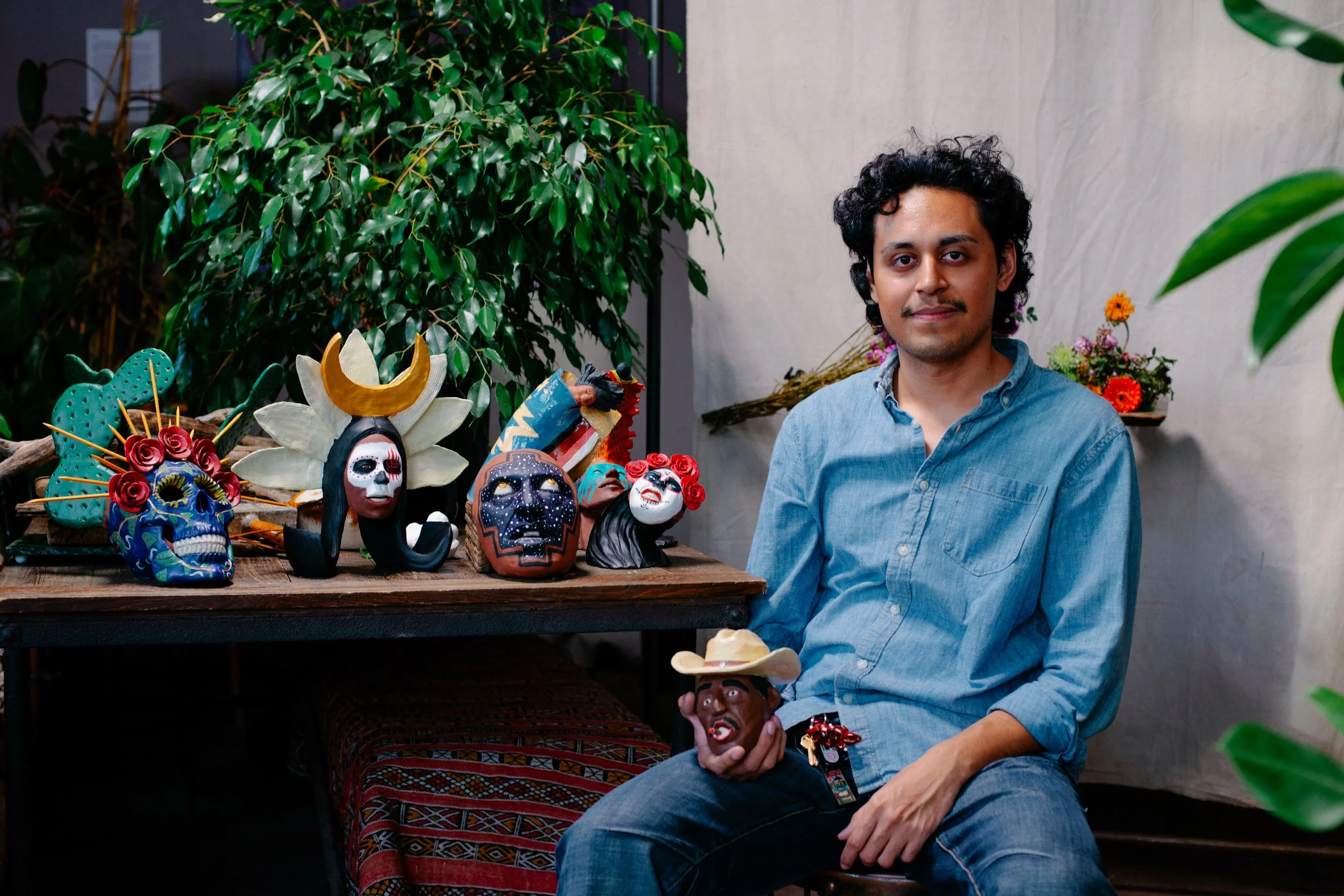 A young man with curly black hair and a light blue shirt sitting indoors near a display of colorful Mexican masks and figurines, with a backdrop of green foliage and a light-colored wall.
