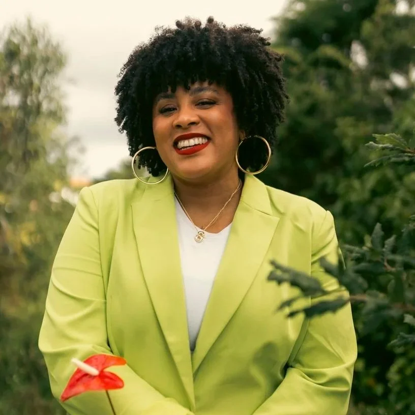 A woman with curly black hair wearing a lime green blazer, white top, and gold hoop earrings, standing outdoors with greenery in the background, smiling with red lipstick.