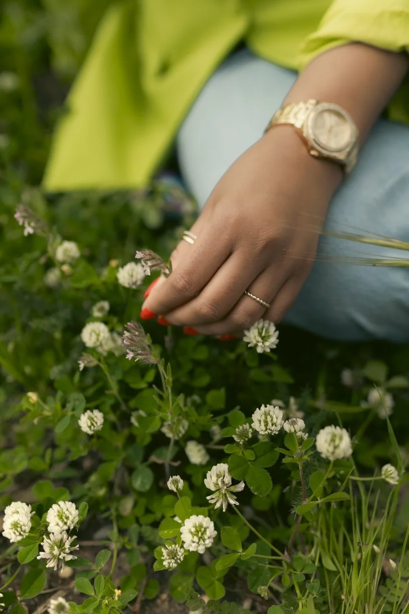Close-up of a person's hand with a gold watch and rings, reaching into a patch of white clover flowers, wearing a yellow jacket and light blue jeans.