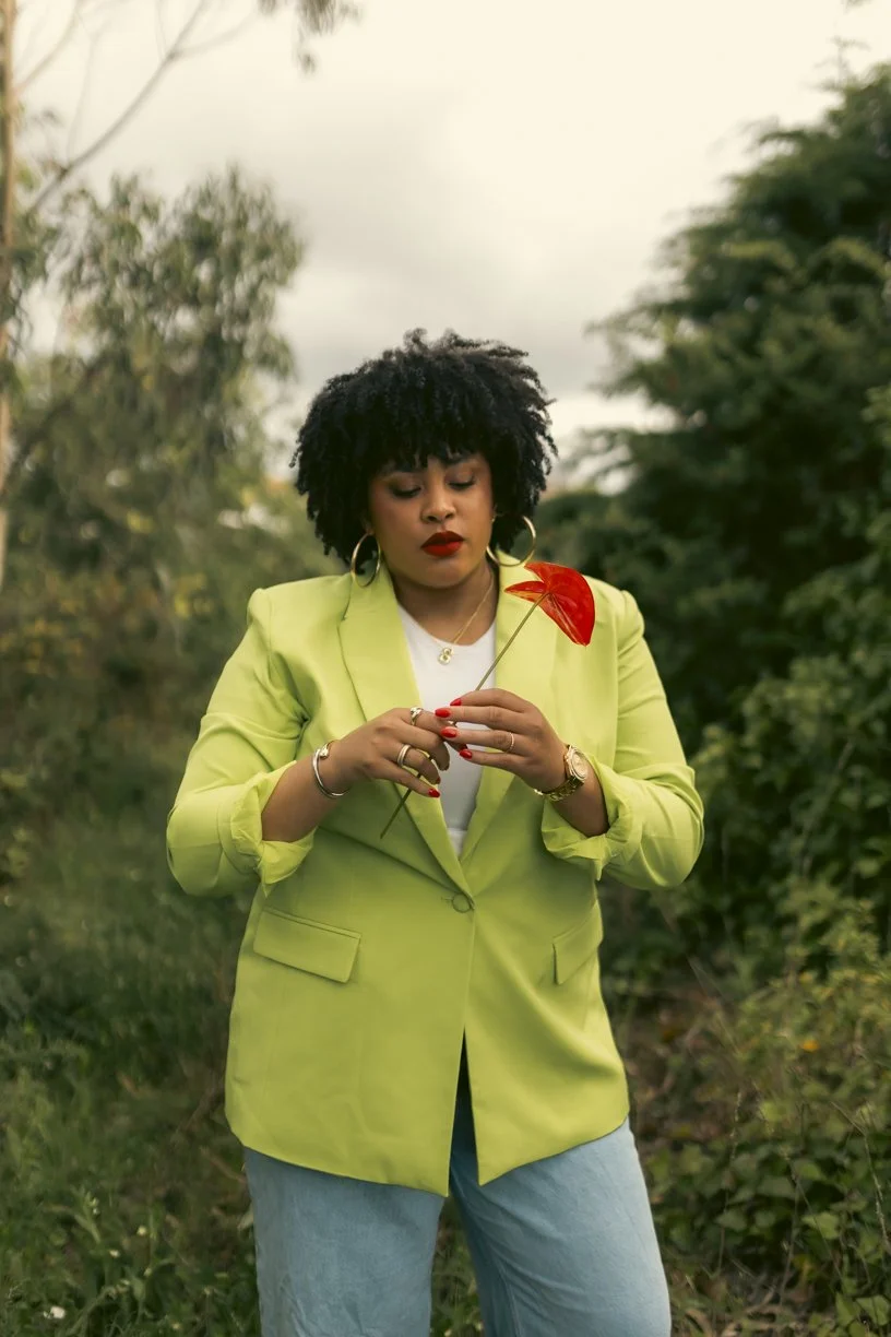 A woman with curly black hair wearing a bright lime green blazer and jeans, holding a red flower outdoors with green trees in the background.