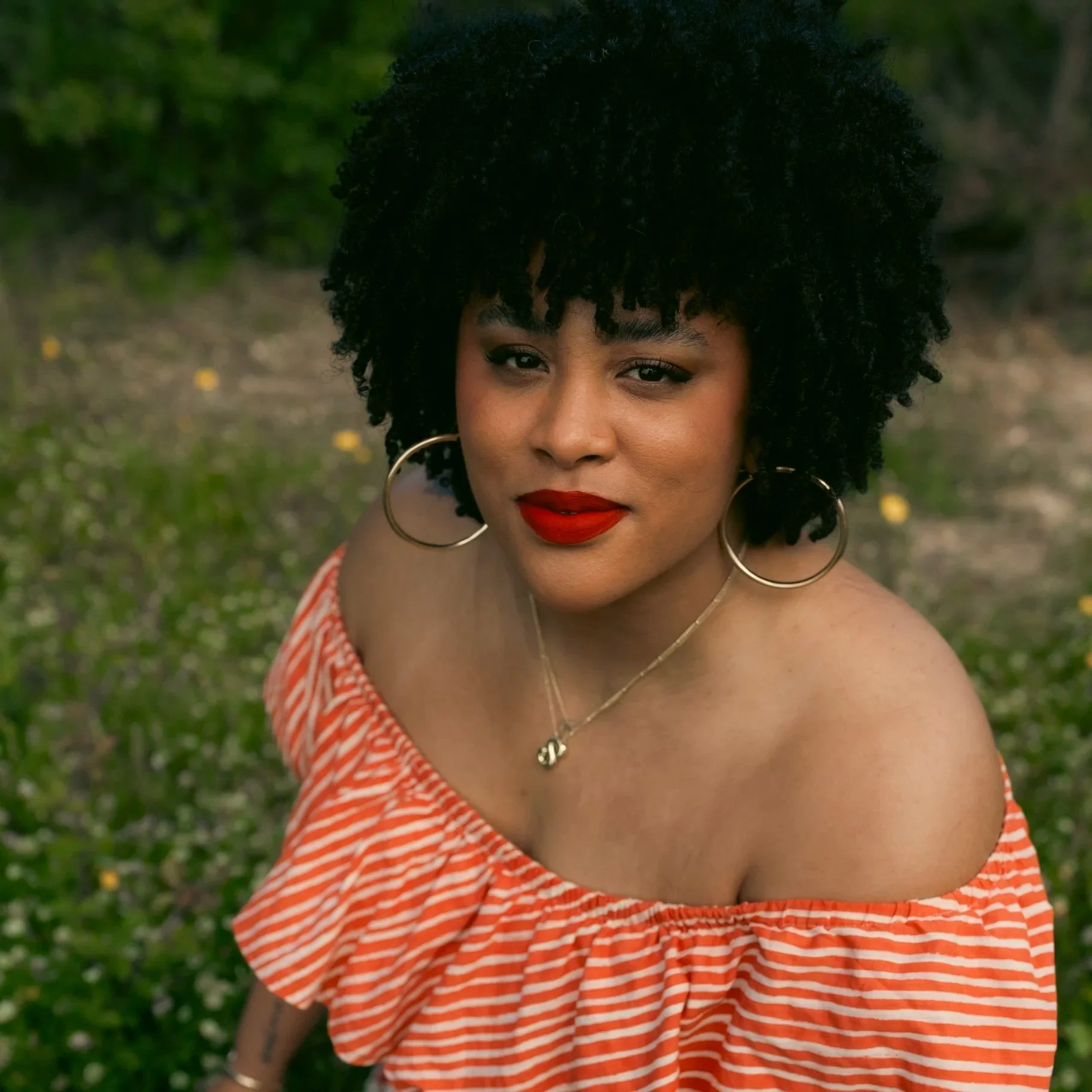 A woman with dark, curly hair, red lipstick, gold hoop earrings, and a layered necklace, wearing an off-the-shoulder orange and white striped top, outdoors with green foliage in the background.