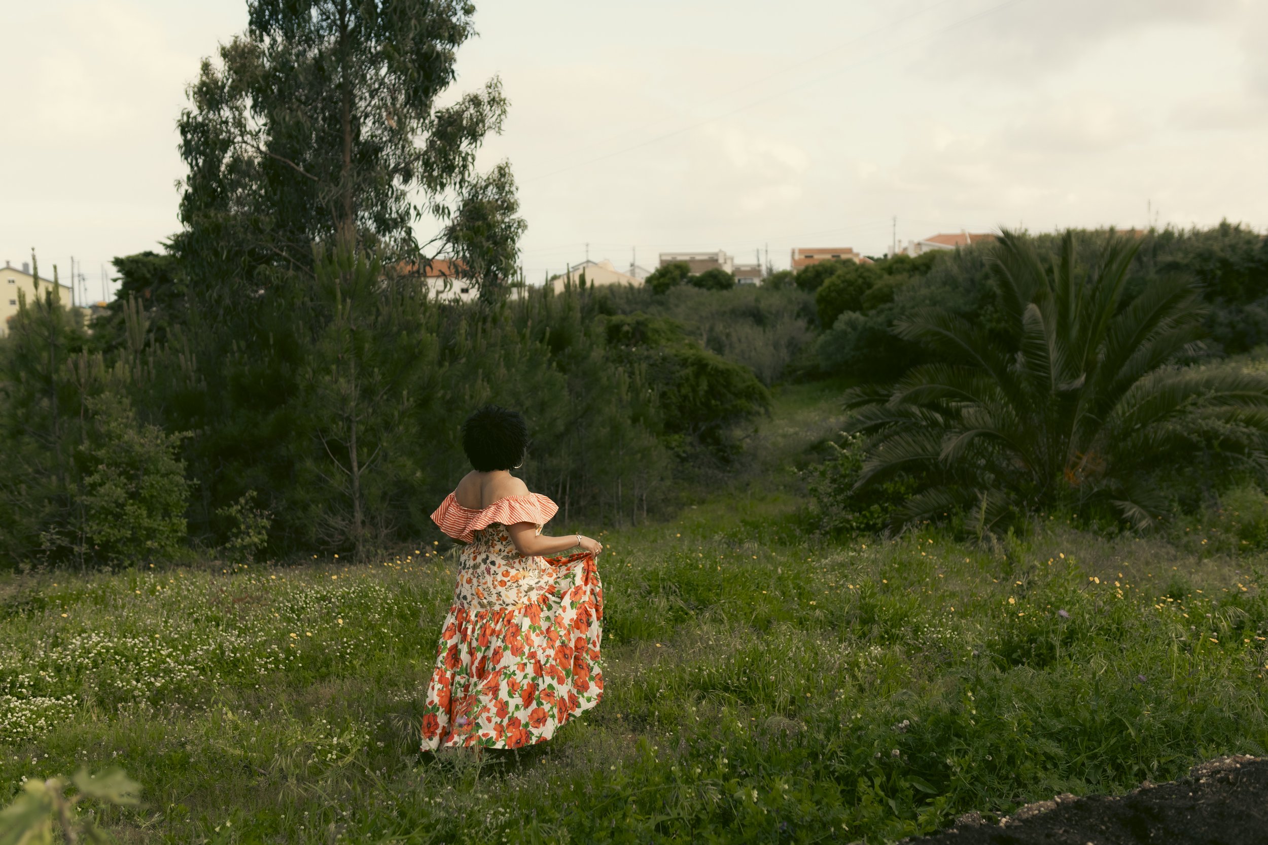 A woman in a floral and striped dress walking through a grassy field with trees and houses in the background.