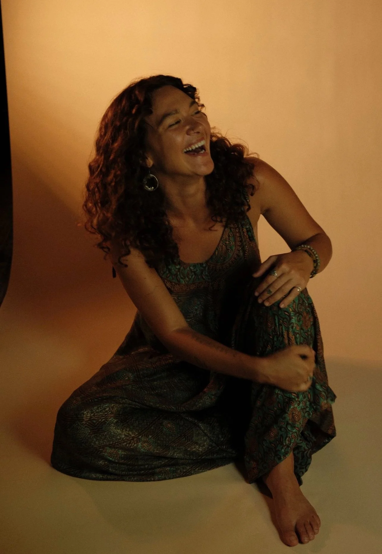 A woman with curly hair laughing and sitting on the floor against a plain beige background, wearing a patterned dress with jewelry.