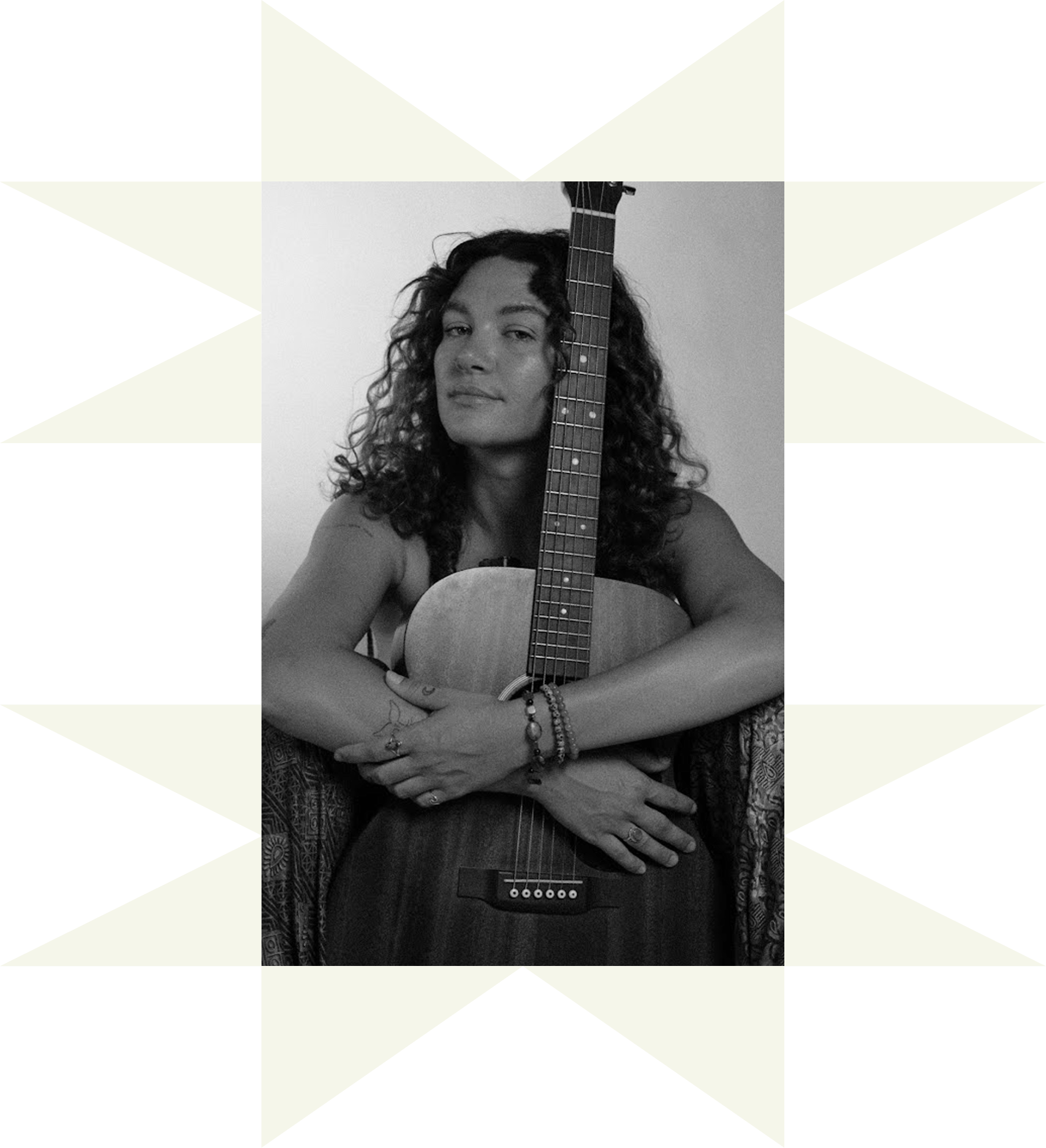A black and white photo of a woman with curly hair, holding an acoustic guitar, looking at the camera, wearing bracelets and rings, in front of a plain background.