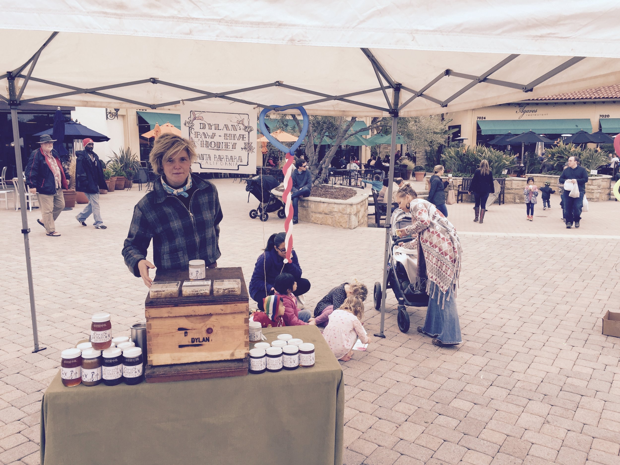 A woman selling jars of honey at an outdoor market stall under a canopy, with several children and adults browsing and walking in the background.