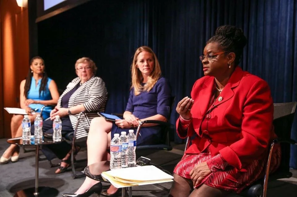 Four women seated on a stage during a panel discussion, with water bottles and documents on small tables in front of them.