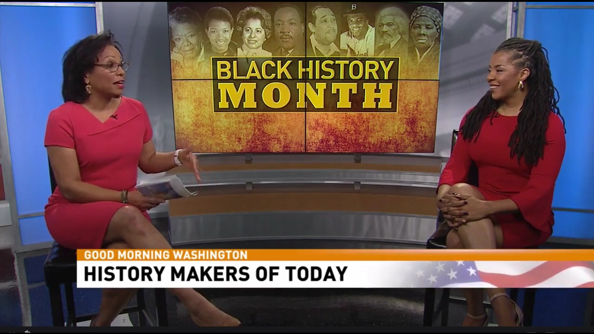 Two women in red dresses are engaged in conversation on a news set. Behind them is a large screen displaying images of diverse Black historical figures and the text 'Black History Month.' A news ticker at the bottom reads 'Good Morning Washington, History Makers of Today.'