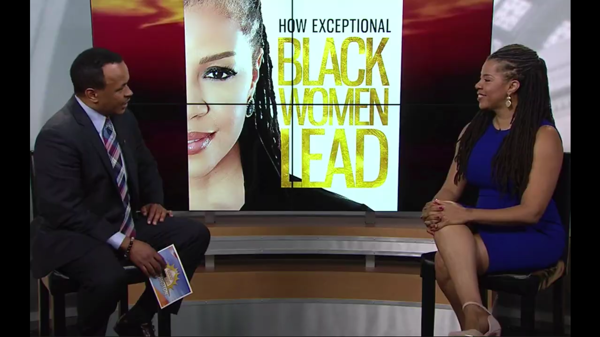 A TV interview with a man and woman in a studio. Behind them is a large screen displaying a woman with dreadlocks and text that reads "How Exceptional Black Women Lead." The man is holding a card with a sun and star design, and the woman is sitting with her hands clasped, smiling.