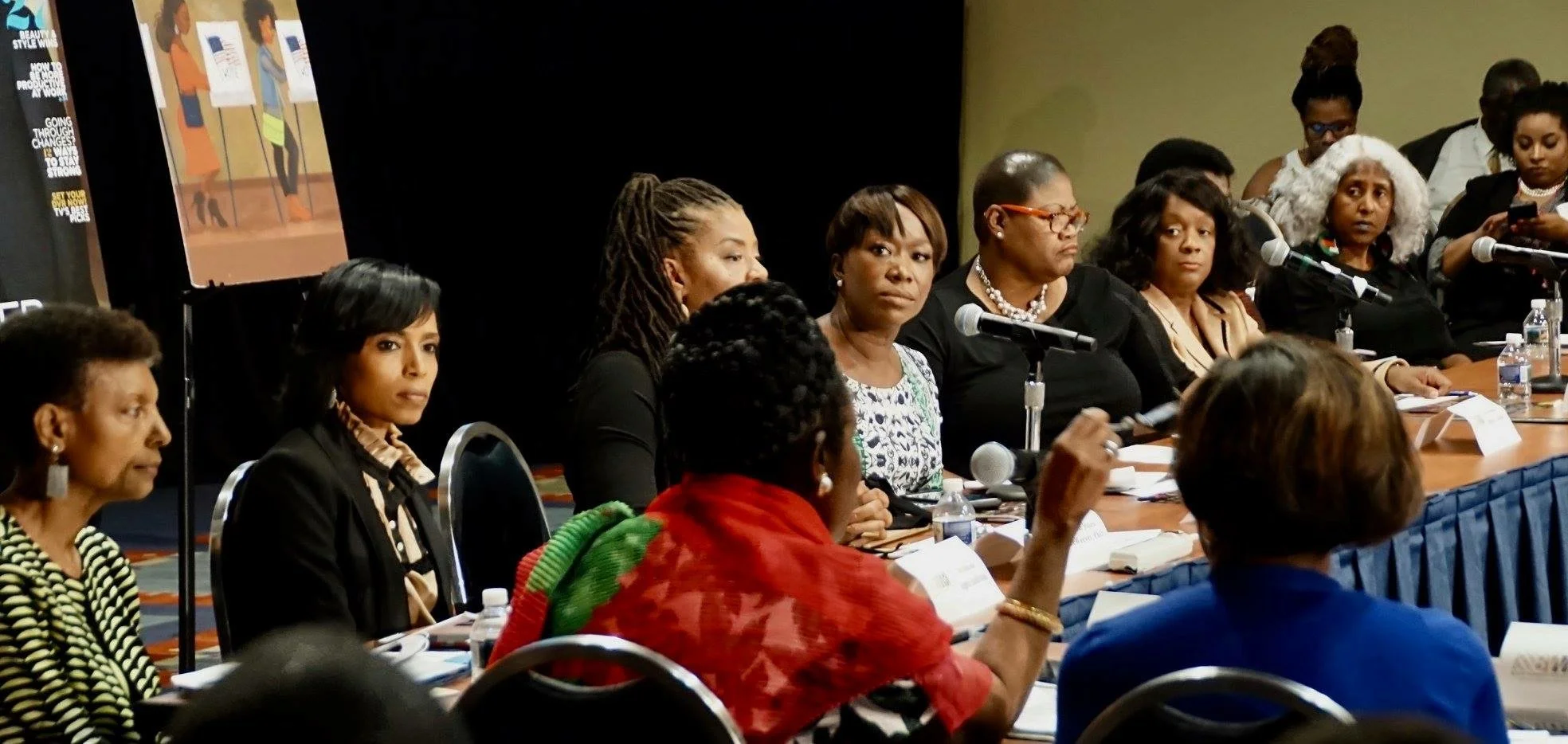 A panel of diverse women seated at a long table during a conference or meeting, with microphones, papers, and water bottles in front of them.