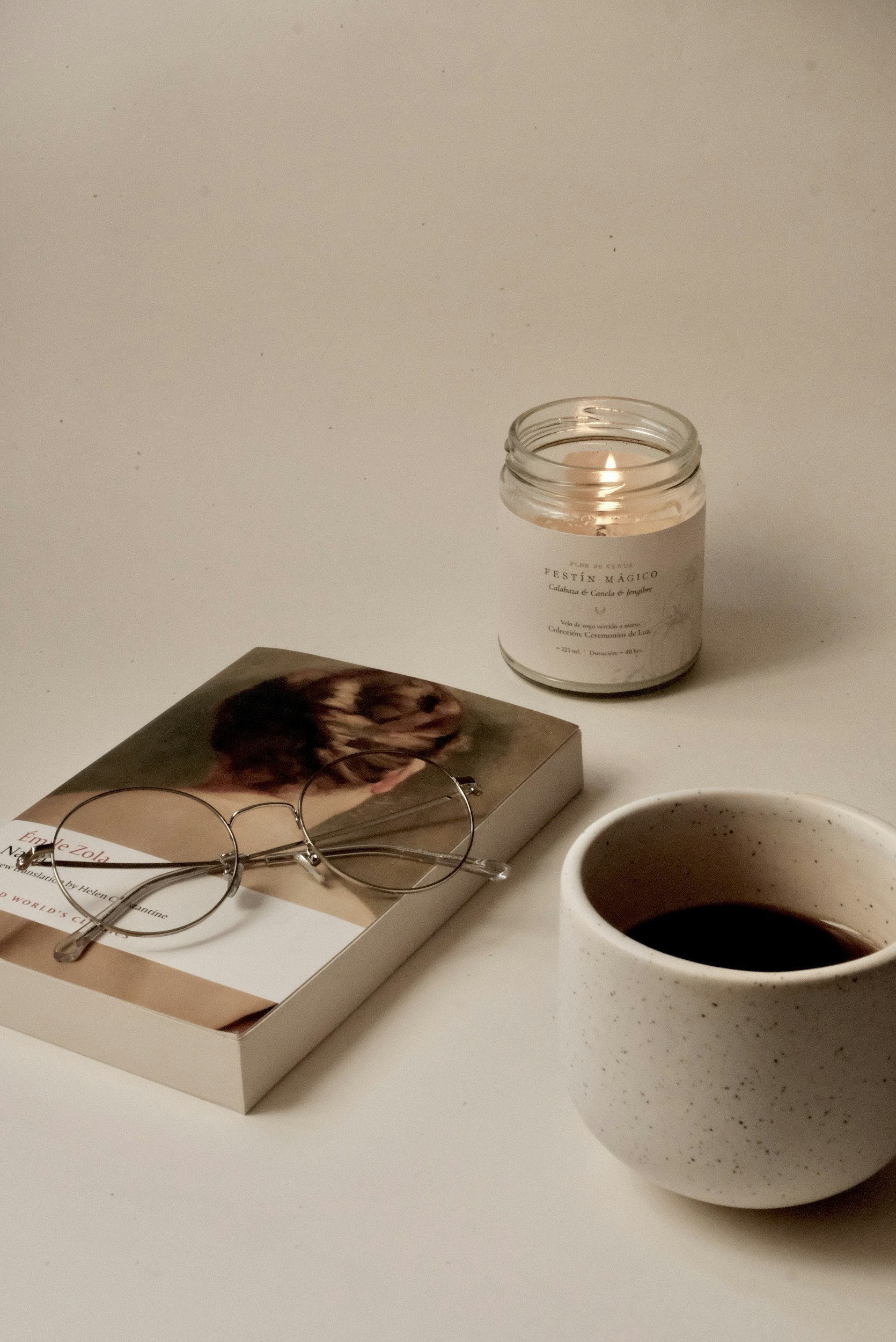 A lit candle in a glass jar, a book with reading glasses resting on top, and a mug filled with black coffee on a white surface against a plain background.