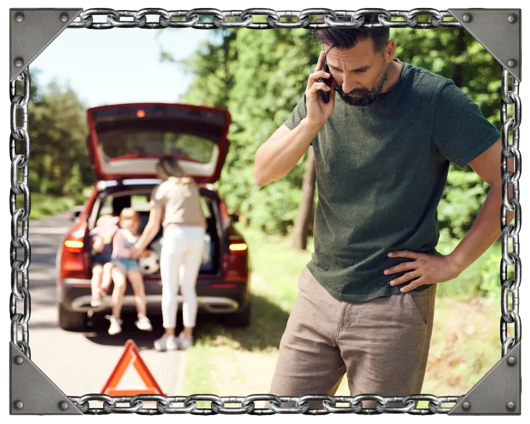 A man standing with a hand on his hip and talking on a cellphone, with a worried expression, on a sunny day near a car with its trunk open, involving two children sitting inside, on a suburban street lined with trees.