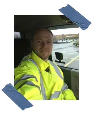 A man in a yellow safety jacket sitting inside a vehicle, smiling at the camera, with a Walmart store visible through the window in the background.