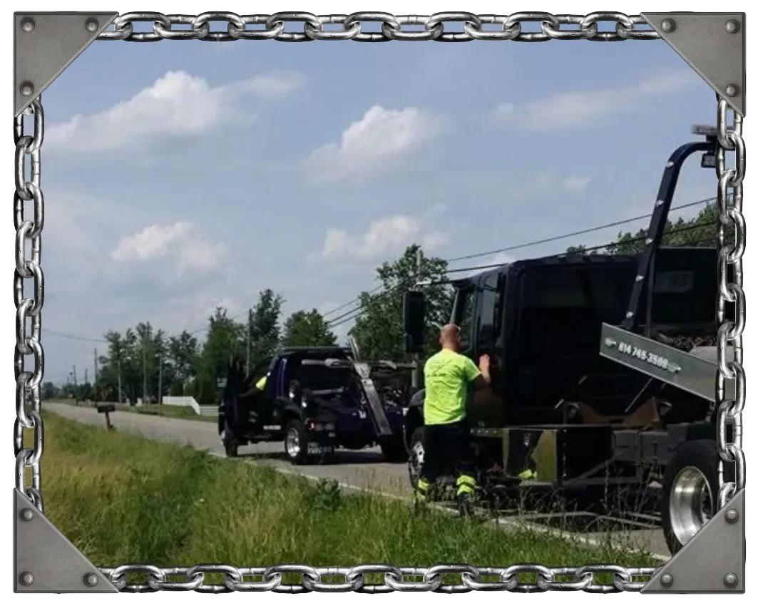 A tow truck assisting a damaged vehicle on the side of a rural road with a person in a neon yellow shirt standing next to the truck.
