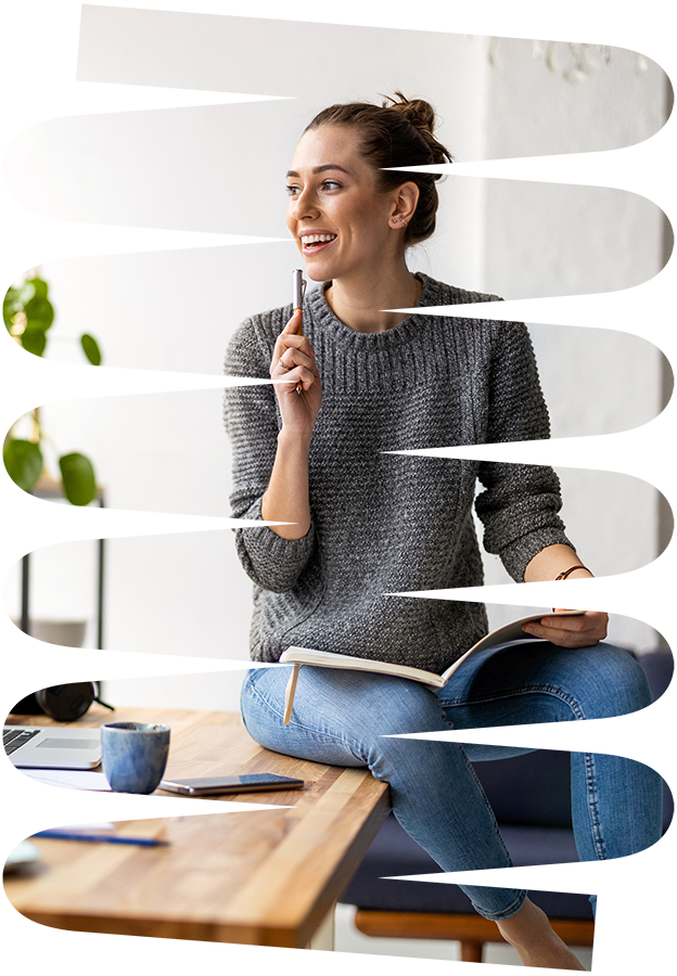 A woman sitting on a desk in a casual gray sweater and blue jeans, smiling and holding a pen near her face while looking to the side.
