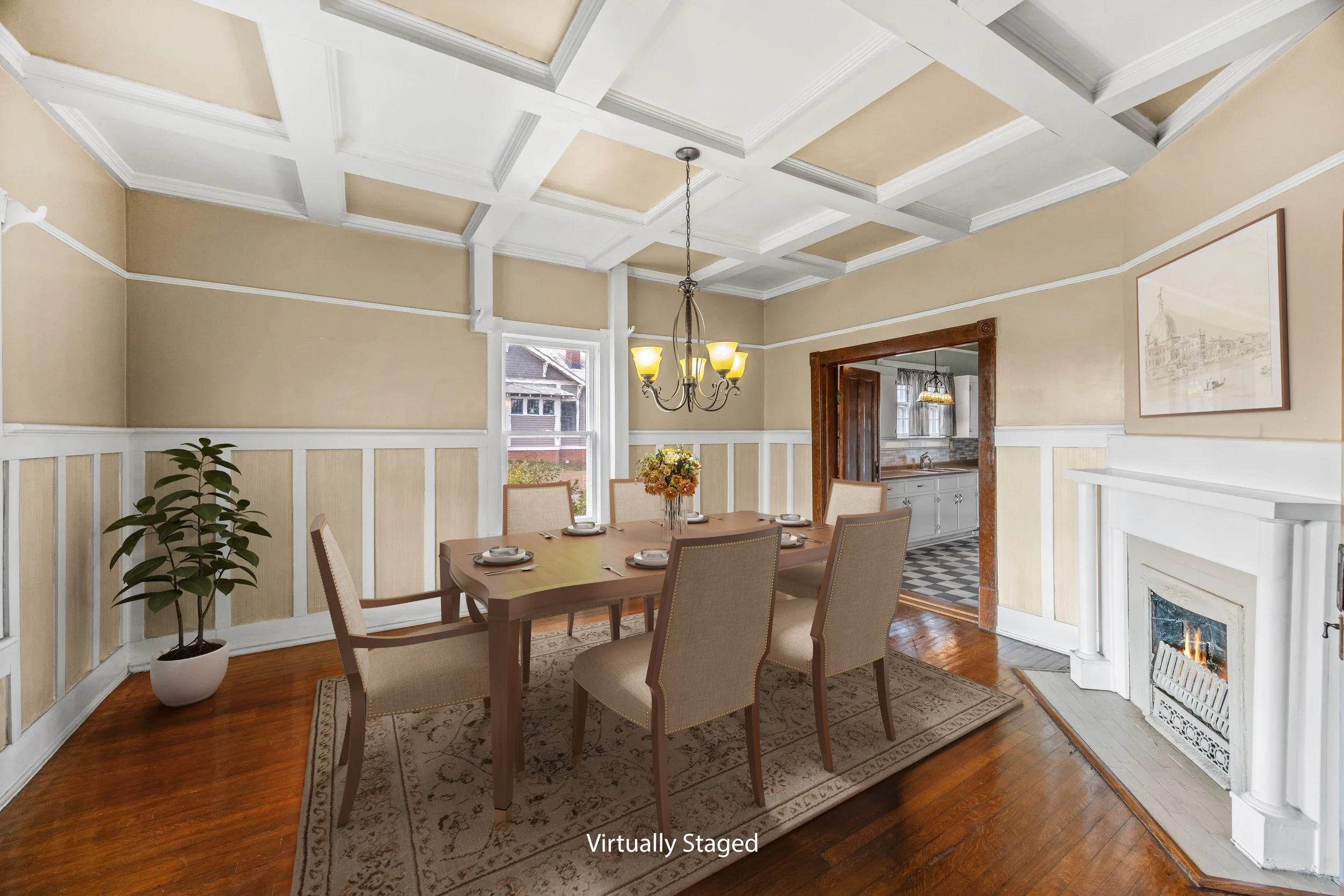 Dining room with a wooden table set with plates and silverware, six beige upholstered chairs, a hanging chandelier, a large window, a potted plant, a patterned rug, and an adjacent kitchen with checkered floor visible through a doorway, and a fireplace in the corner.