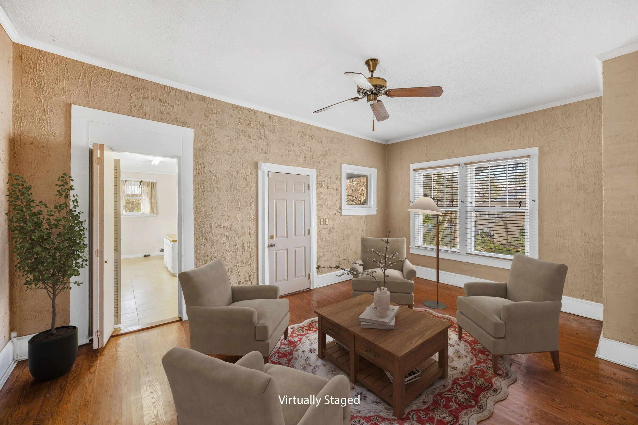 Living room with beige textured walls, hardwood flooring, four beige armchairs around a wooden coffee table on a patterned rug, a floor lamp, windows with blinds, a ceiling fan, potted plant, and a doorway leading to another room.