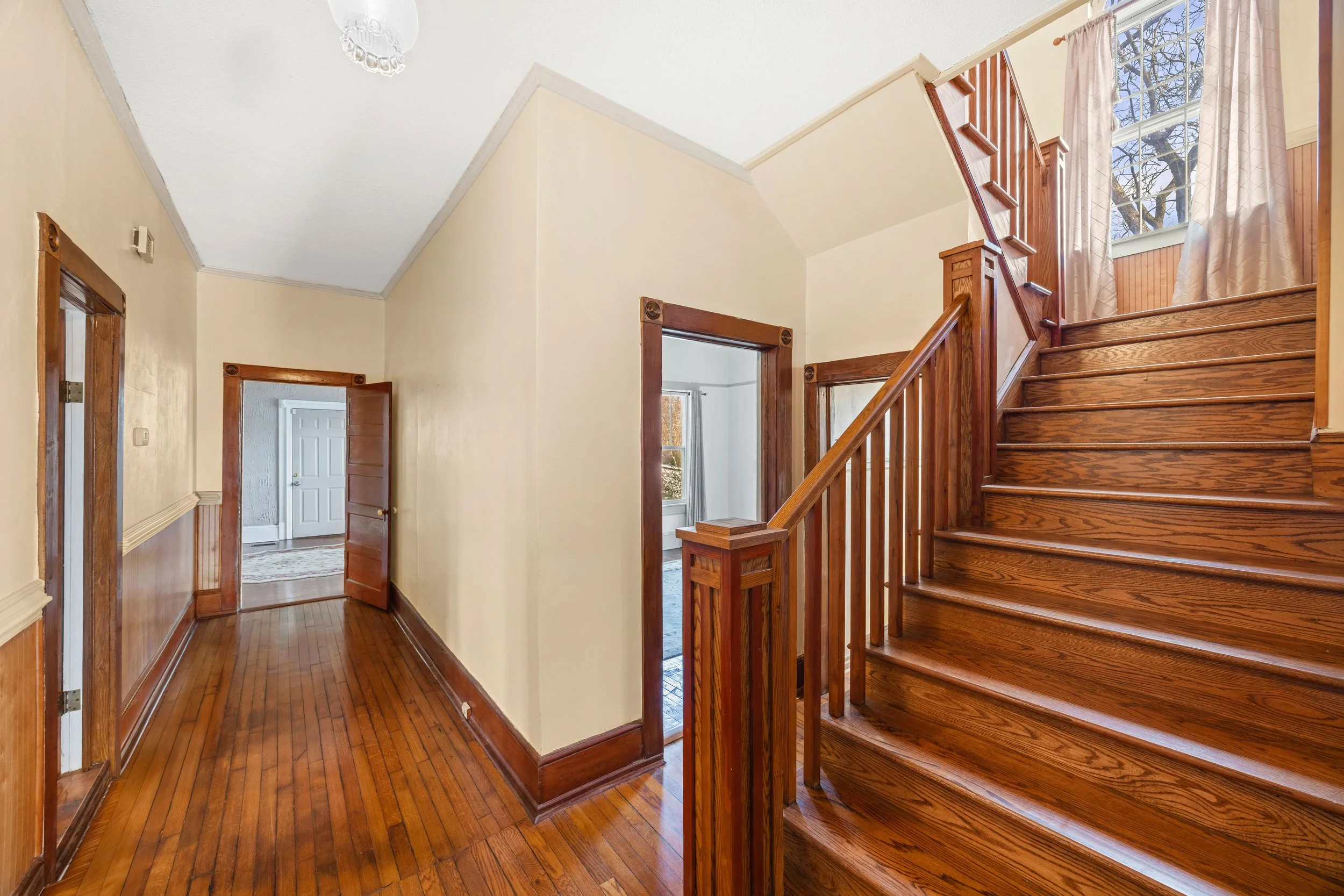 Interior view of a house hallway with hardwood floors, a staircase with wooden handrails, and multiple doorways leading to other rooms, with a large window with curtains at the top of the stairs.