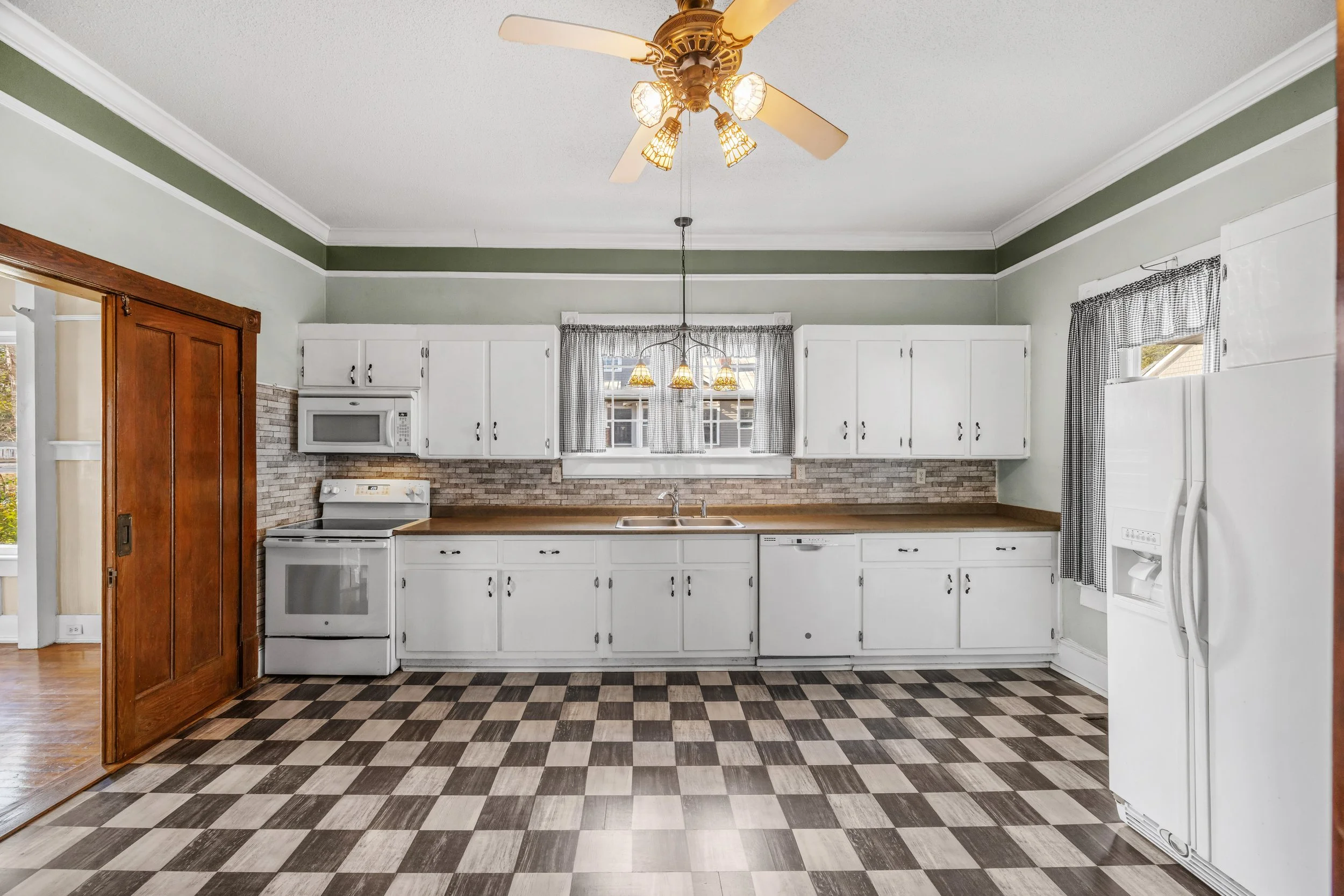 Kitchen with white cabinets, a brick backsplash, and a checkered floor. Includes a white refrigerator on the right, an electric stove, microwave, sink under a window, and a wooden sliding door on the left.