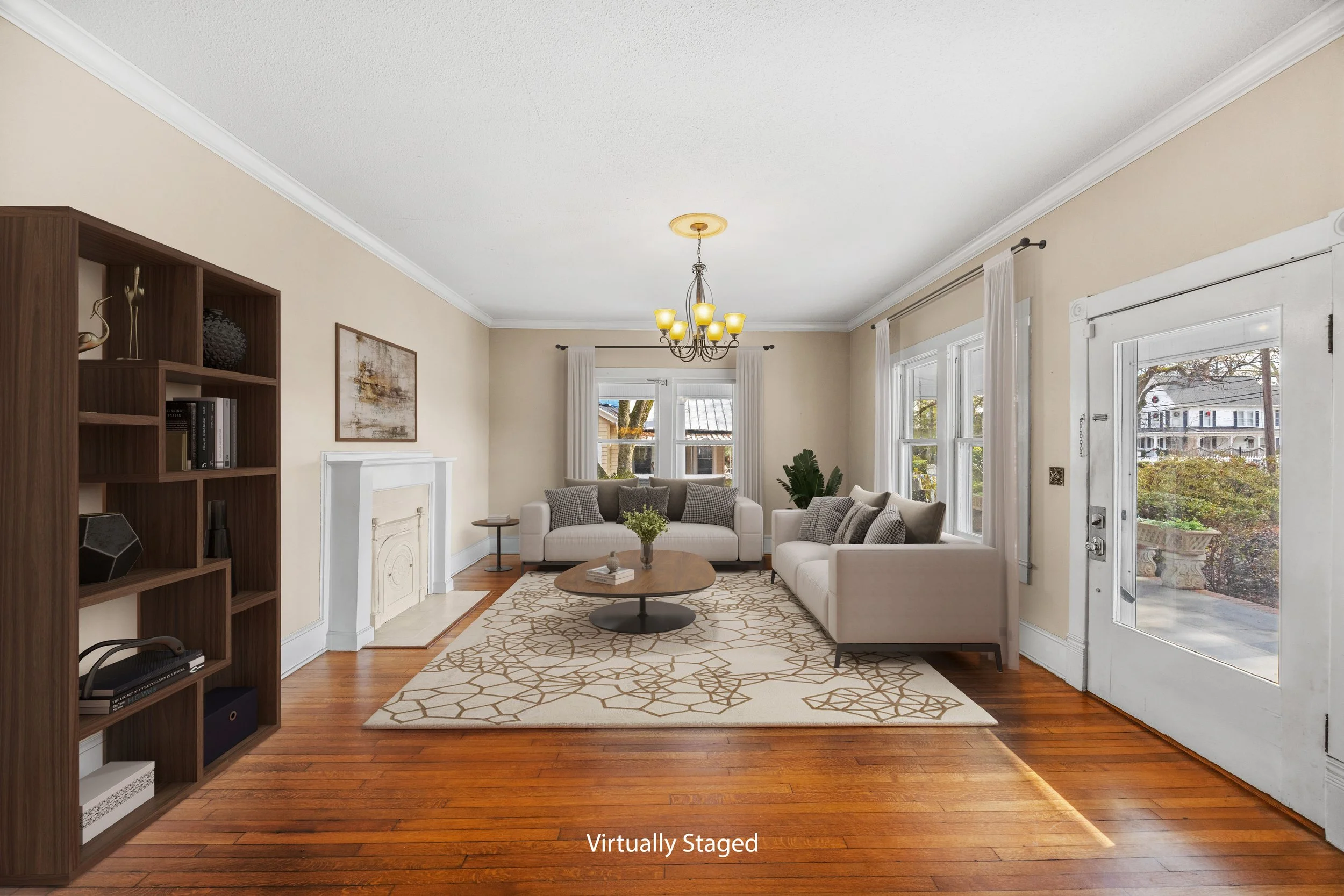 A spacious living room with hardwood floors, two white sofas, a round wooden coffee table with books and a plant, a patterned area rug, a dark wood bookshelf, a white fireplace mantel, large windows with white curtains, a glass door, and a chandelier hanging from the ceiling.