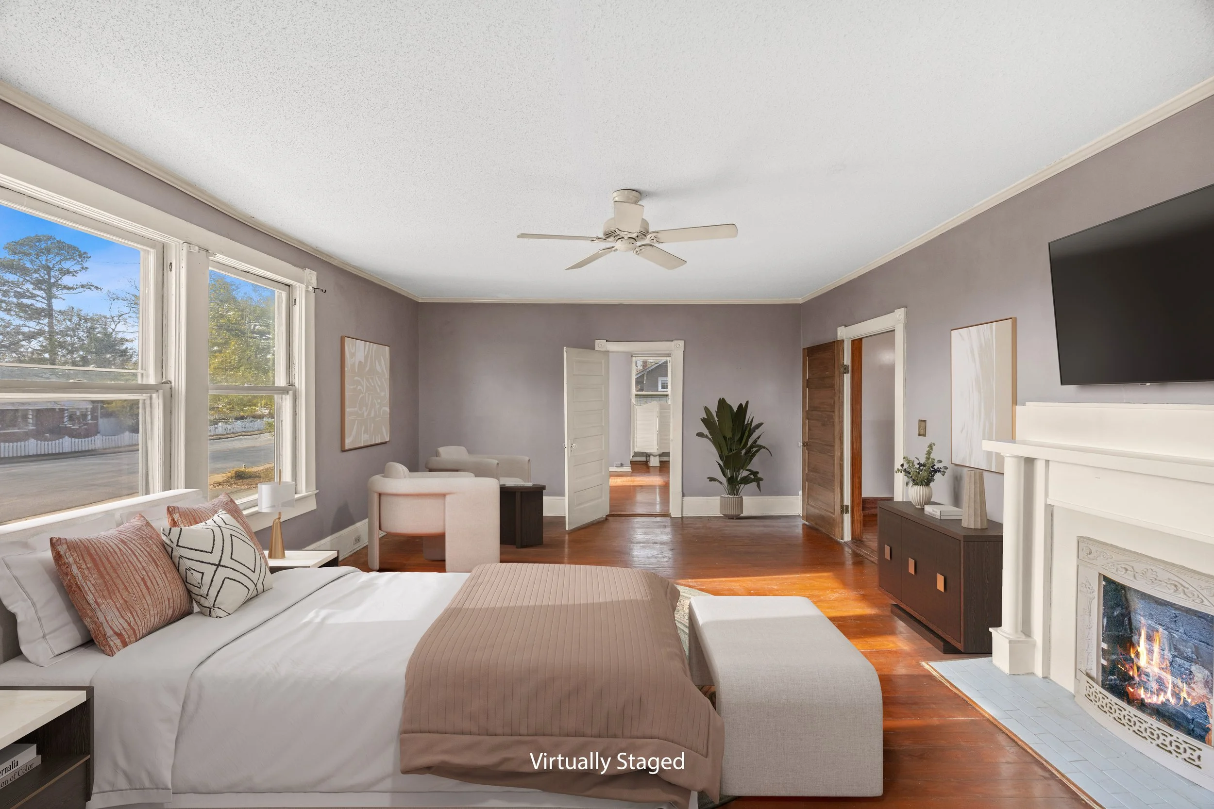 A bedroom with mauve walls, hardwood floors, large windows, a white bed with patterned pillows, a fireplace, a mounted TV, and minimal decor.