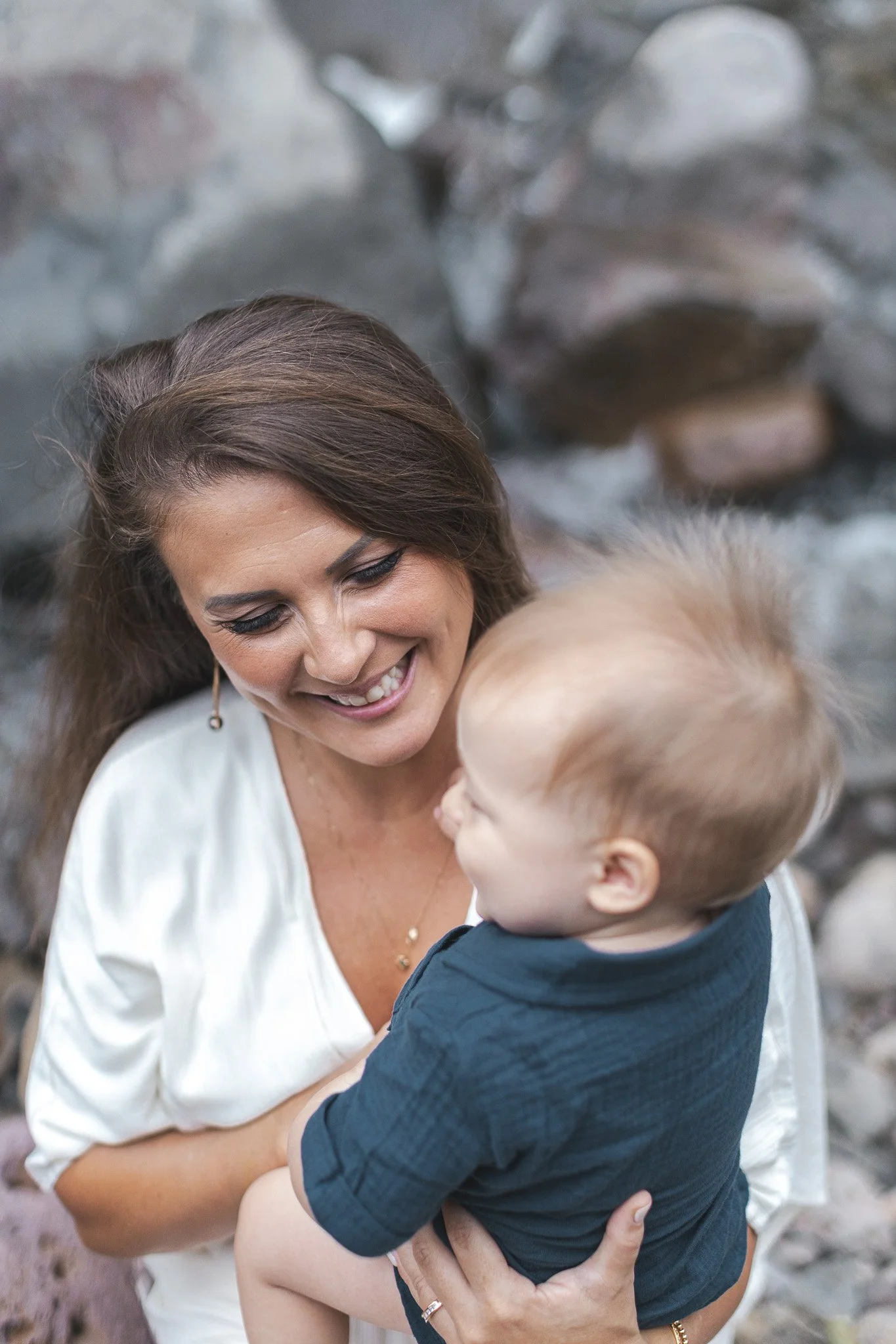 A woman with long brown hair and a white top smiling and holding a young boy with light hair and a navy shirt, with a blurred rocky background.