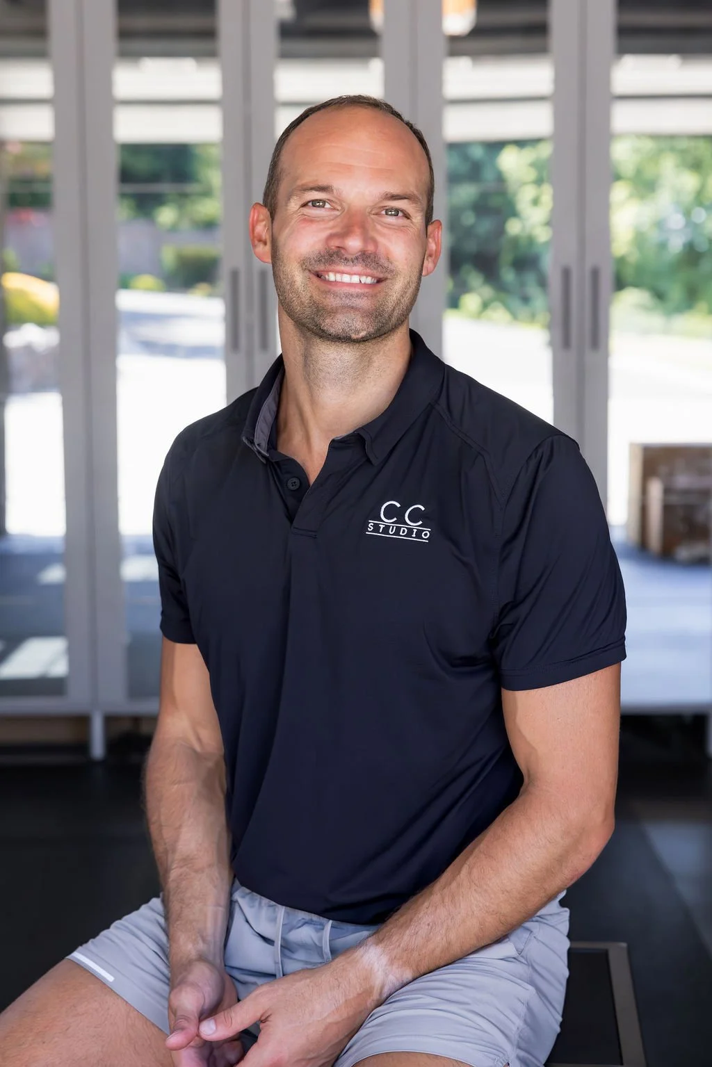 A smiling man with a beard and light skin wearing a navy blue polo shirt with 'CC Studio' logo, sitting indoors with glass doors and greenery outside behind him.